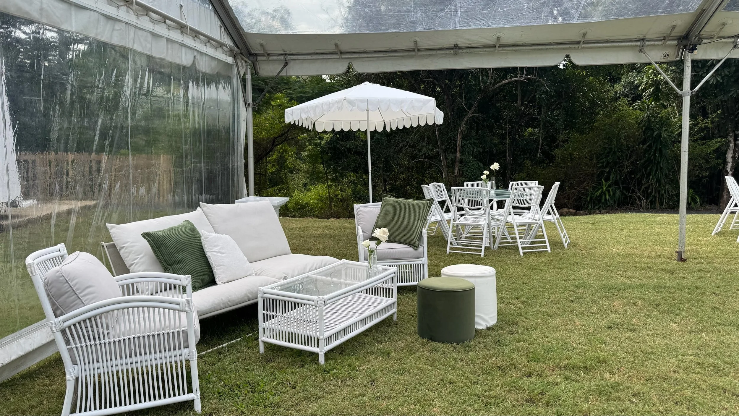 Outdoor patio with white wicker furniture, green and white pillows, glass coffee table, white chairs and round table with chairs under a white patio umbrella, grassy area with trees in the background.