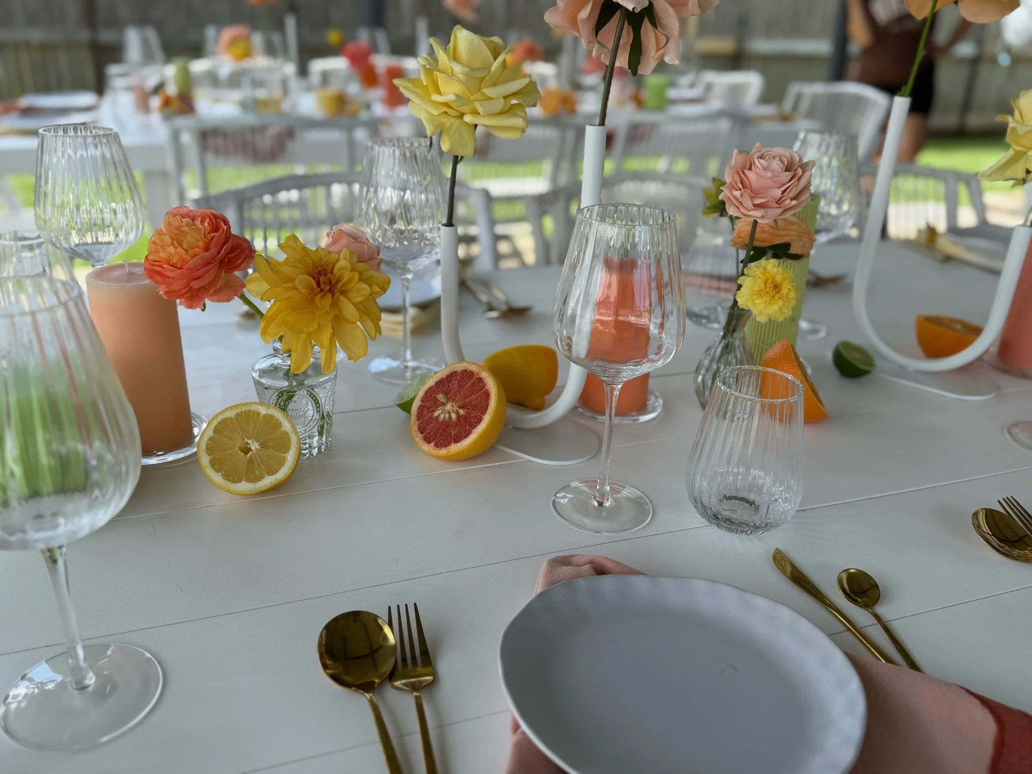 Decorated table with colorful flowers in vases, sliced citrus fruits, wine glasses, and gold utensils for a celebration or event.