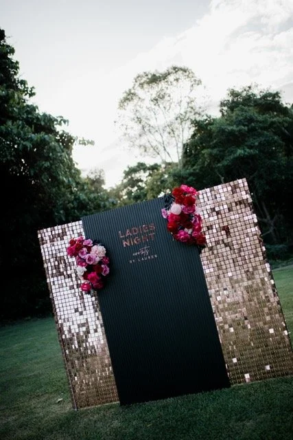 Outdoor event setup with a decorated stand covered in pink and red flowers, featuring a black central panel with 