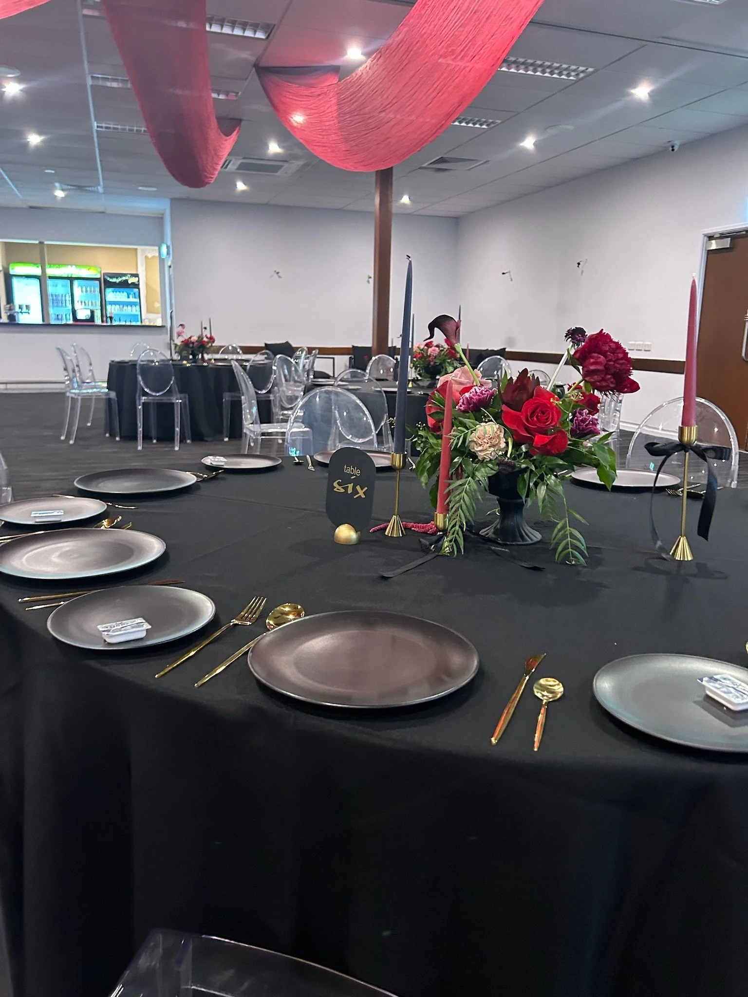 A banquet table decorated with a black tablecloth, a flower centerpiece with pink, red, and purple flowers, and tall candles in gold holders. The table is set with black and gold cutlery, gray plates, and clear chairs. In the background, there are ad