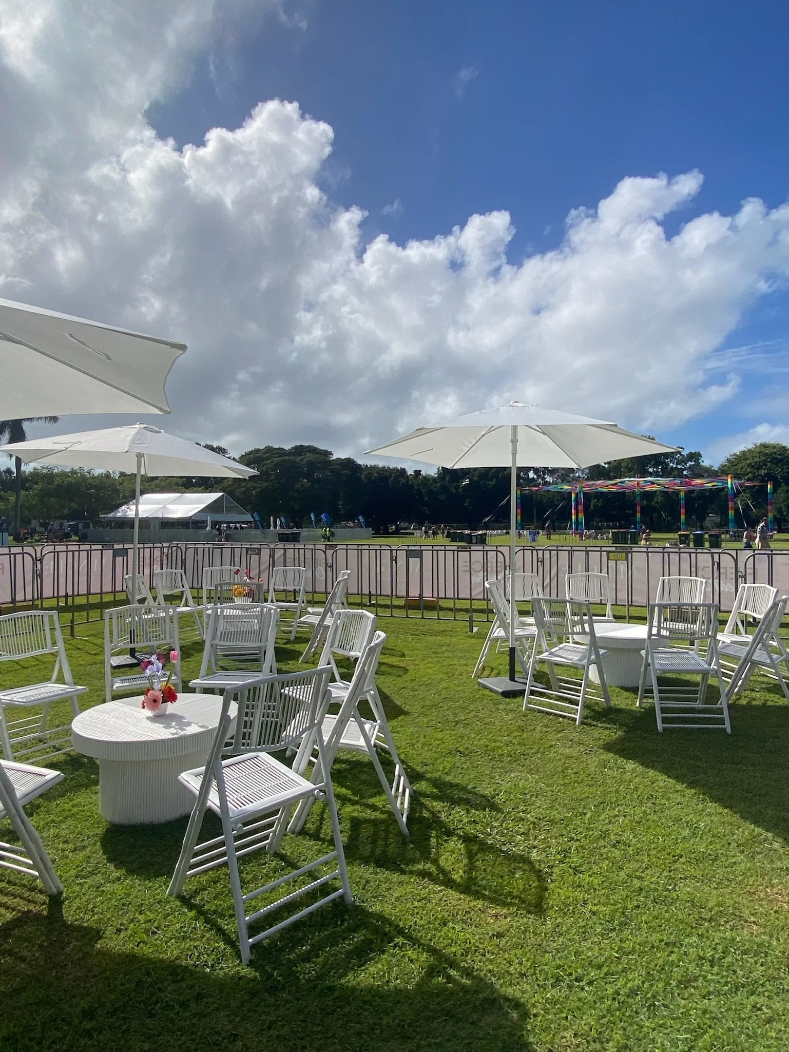 Outdoor event setup with white chairs, round tables, and large white umbrellas on a grassy field under a partly cloudy sky.