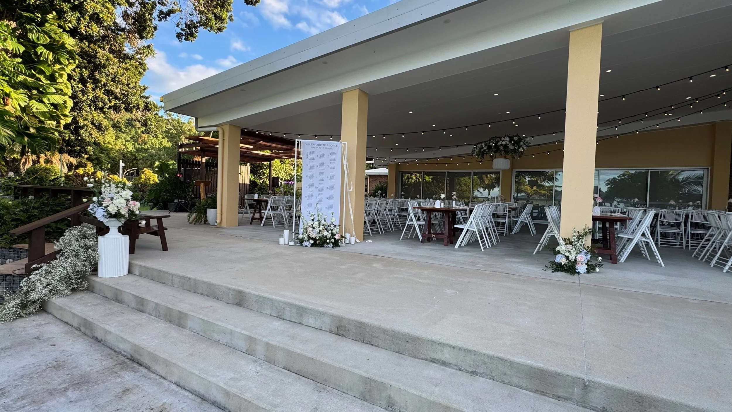 Outdoor event space with tables and chairs, floral decorations, and string lights under a covered patio, surrounded by greenery.