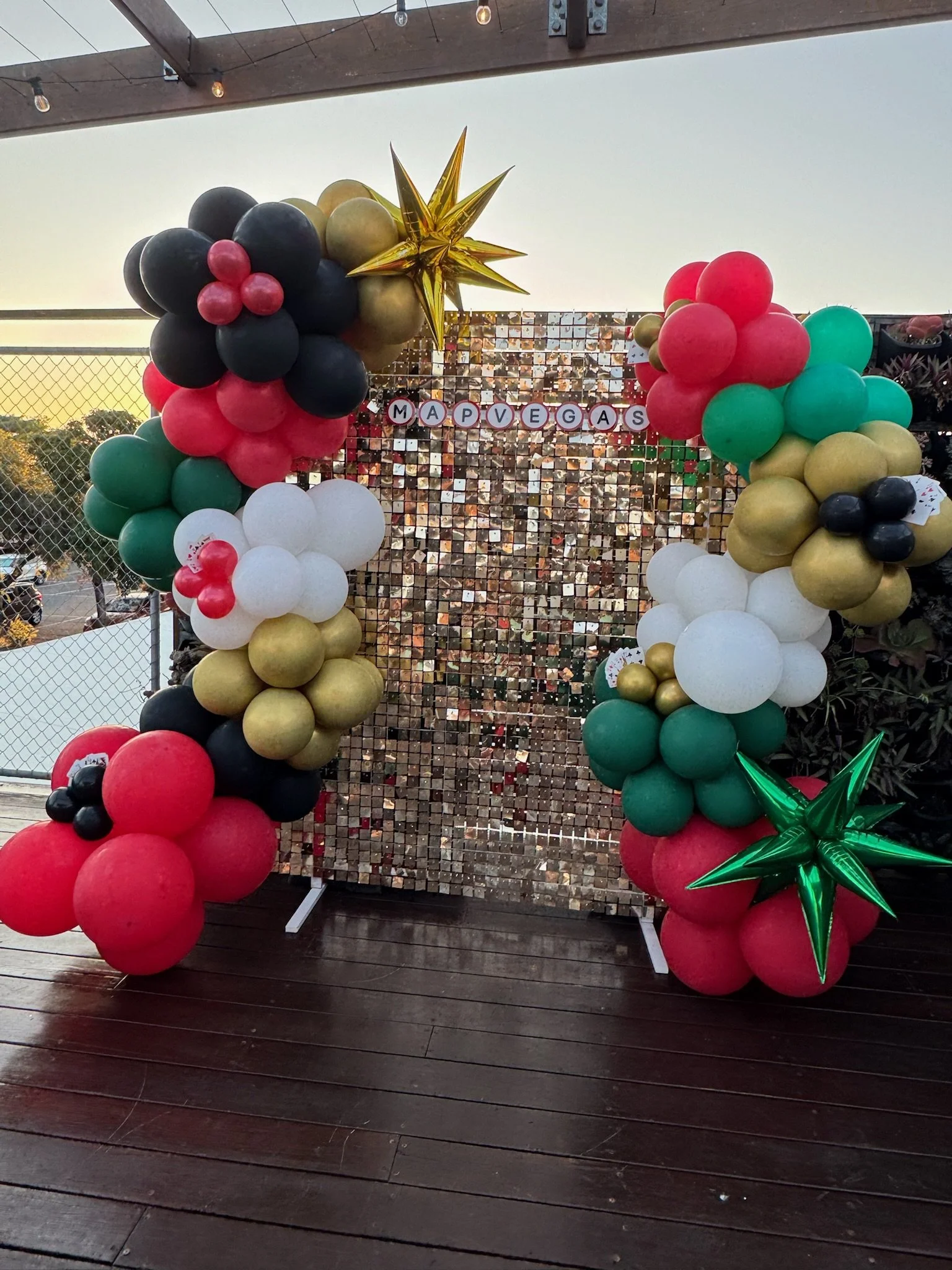 Colorful balloon arch with red, green, black, white, and gold balloons, decorated with gold star and a smaller green balloon star, set against a shiny mosaic backdrop with the words 'MAY PEGAS' on it, on a wooden deck outdoor setting.