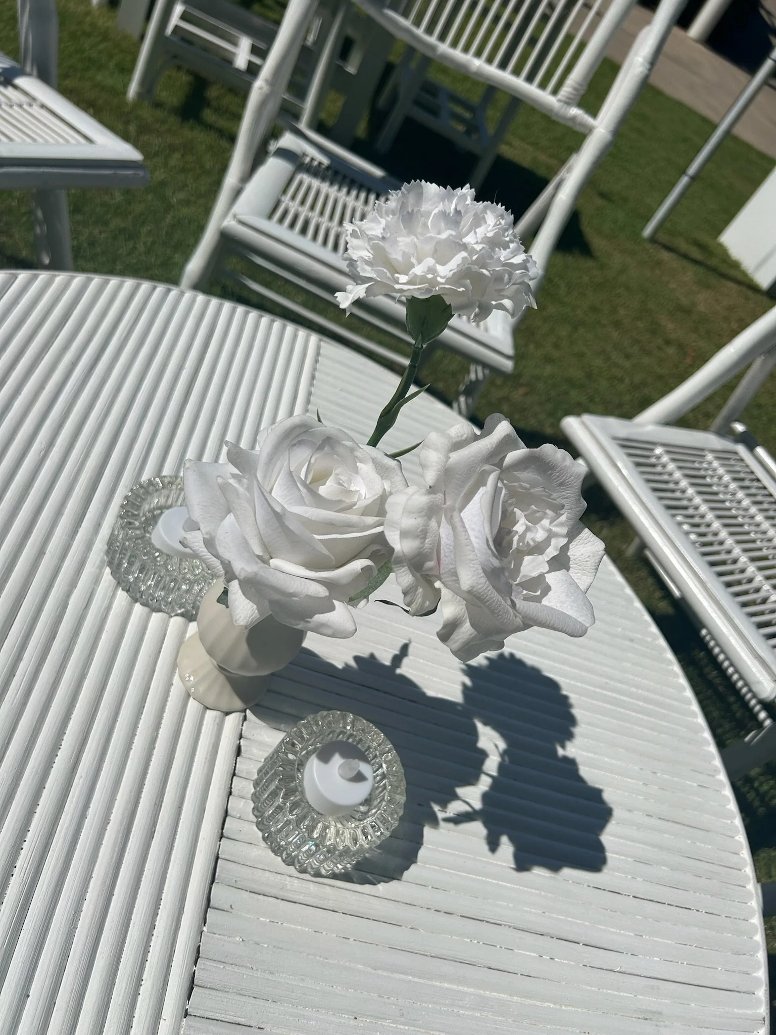 White artificial flowers in a small white vase on a white slatted outdoor table, with glass candle holders, set outdoors with white chairs and a grassy area in the background.