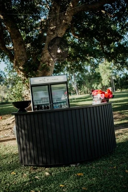 Outdoor black bar with a mini refrigerator, a flower arrangement, and a black bowl, set under a tree in a grassy area.