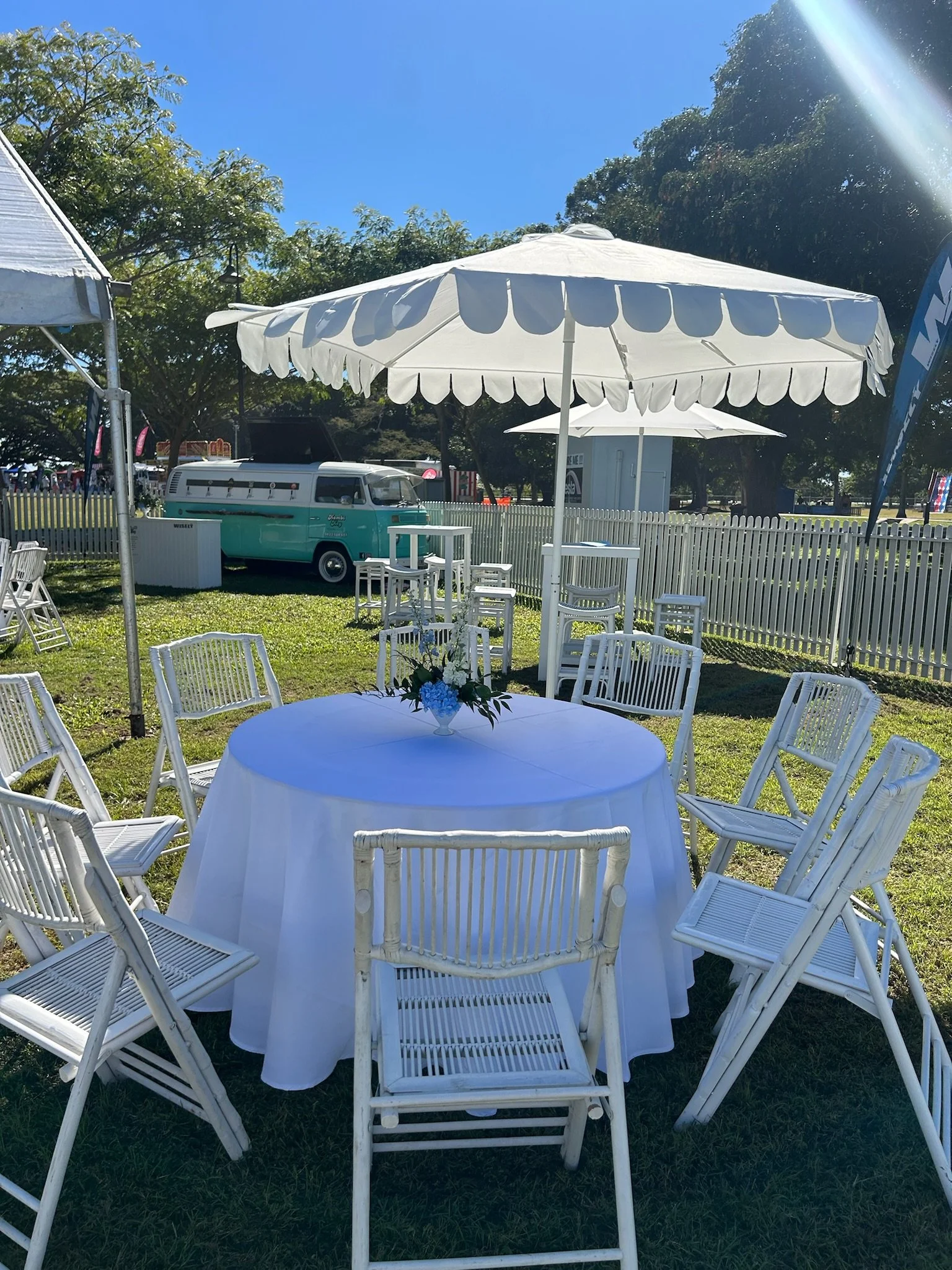 Outdoor event setup with a round table covered in a white tablecloth, surrounded by white chairs, with a large white umbrella overhead, a vintage teal van in the background, and trees under a sunny sky.