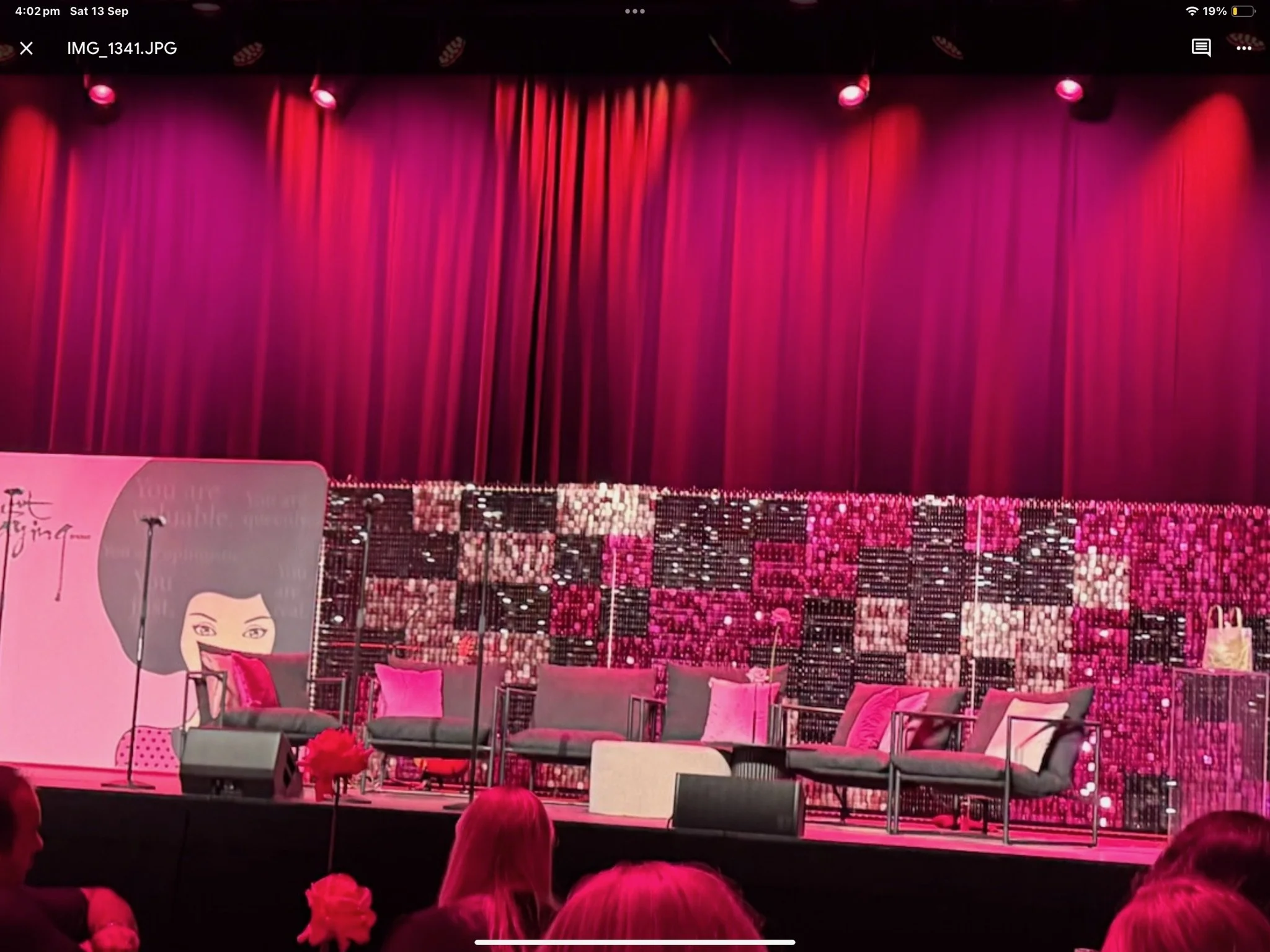Stage with pink lighting, red curtains, and an arrangement of chairs and cushions, with a colorful backdrop and a pink flower in the foreground.