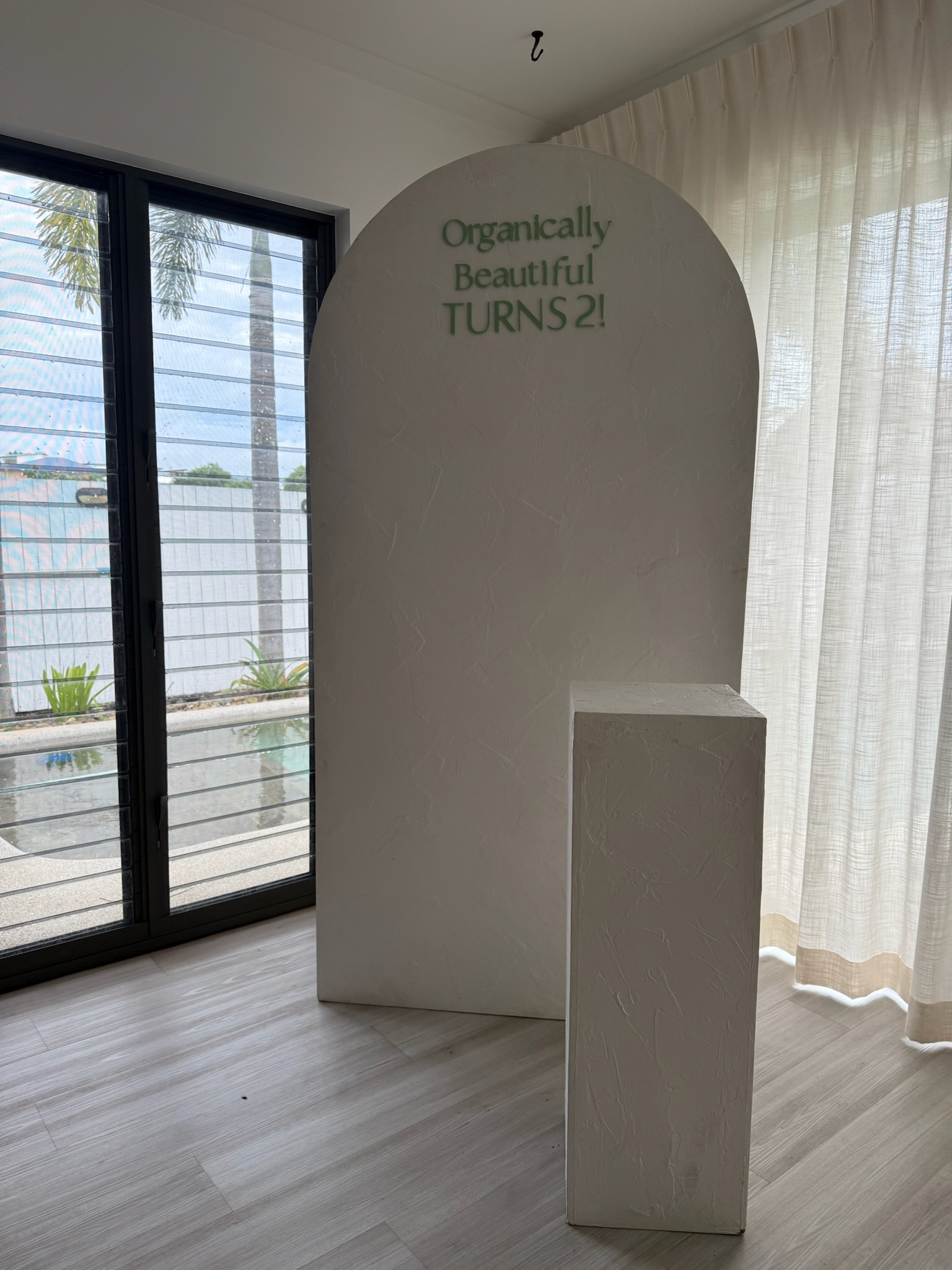 A freestanding display with a rounded top and a rectangular base, with green text that reads "Organically Beautiful TURNS 2!" next to large glass doors with blinds, overlooking a patio with plants and palm trees.