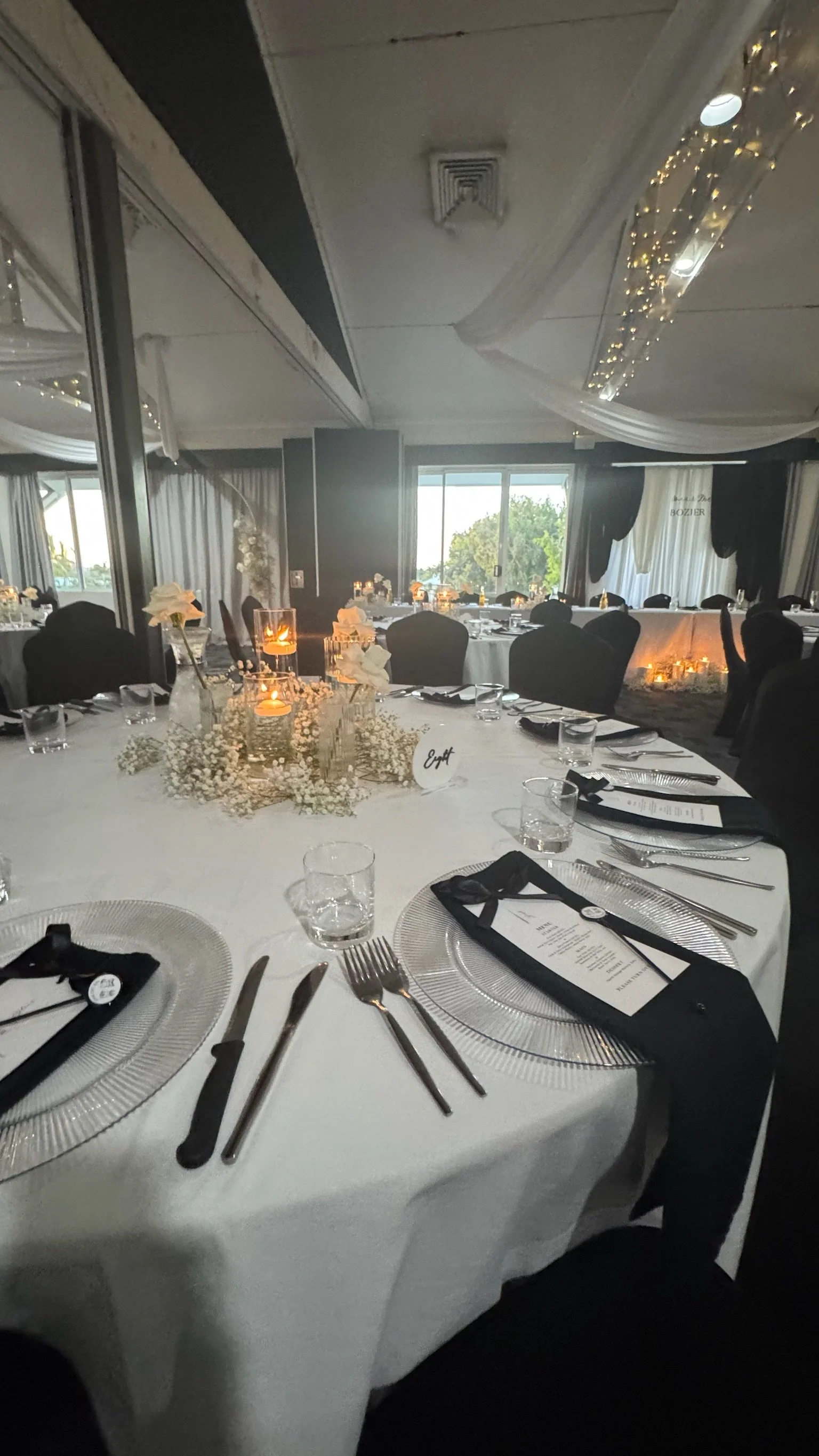 Set dinner table in a decorated banquet hall with black chairs, white tablecloth, floral centerpiece, candles, menus, and place settings with knives, forks, and glasses.