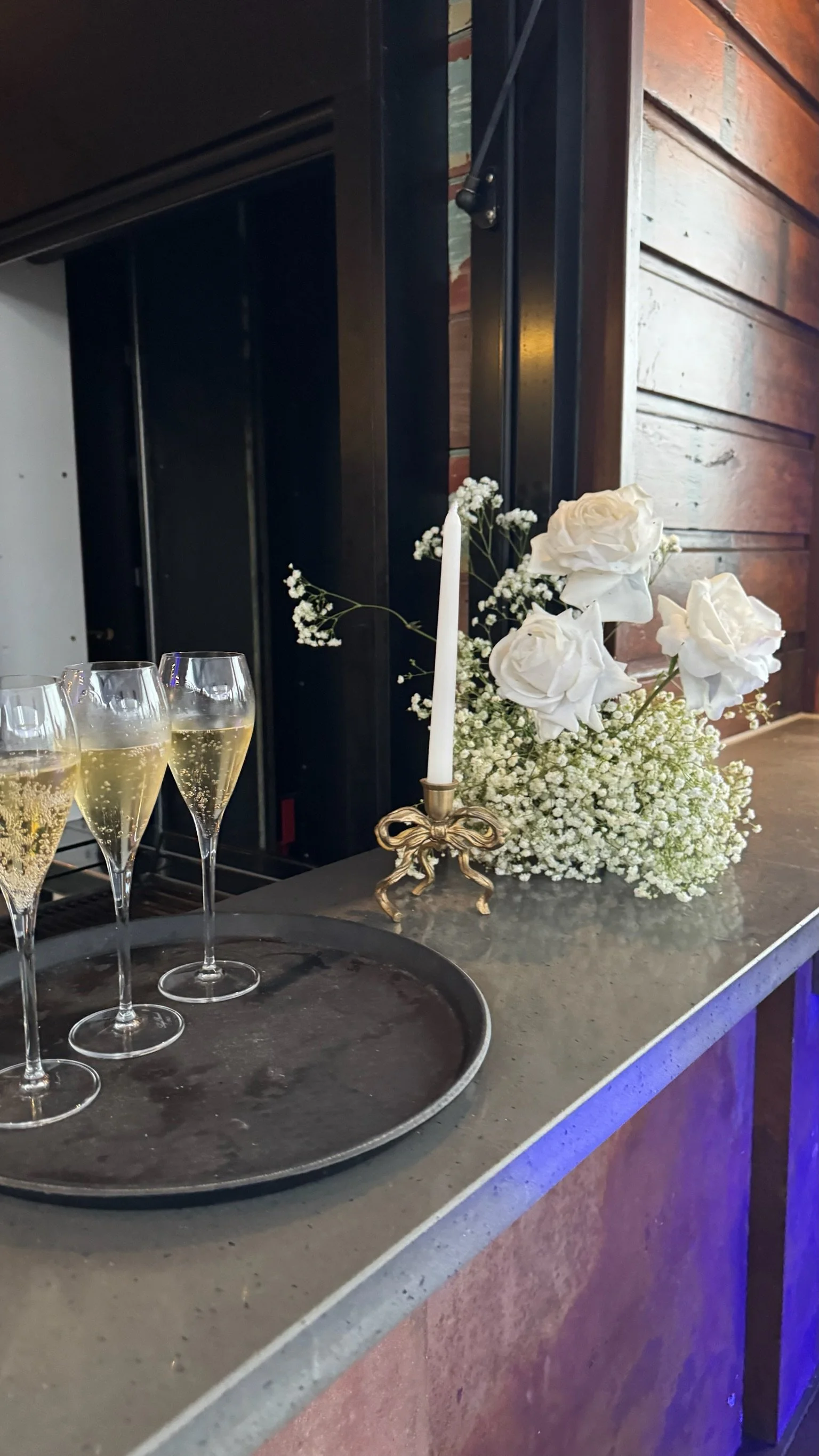 A tray with four glasses of champagne, a white floral arrangement with roses and baby's breath, and a white candle in an ornate gold holder on a gray countertop.
