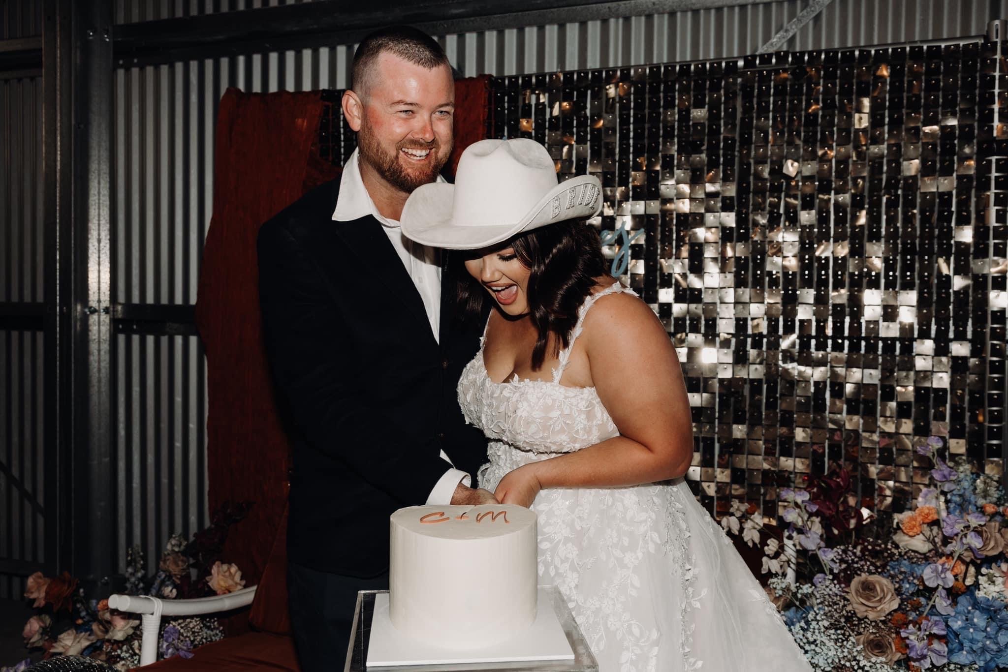 Bride and groom cutting wedding cake, the bride wearing a white lace wedding gown and a white cowboy hat, the groom in a black suit with a white shirt, smiling, with decorative flowers and a sparkling backdrop in the background.