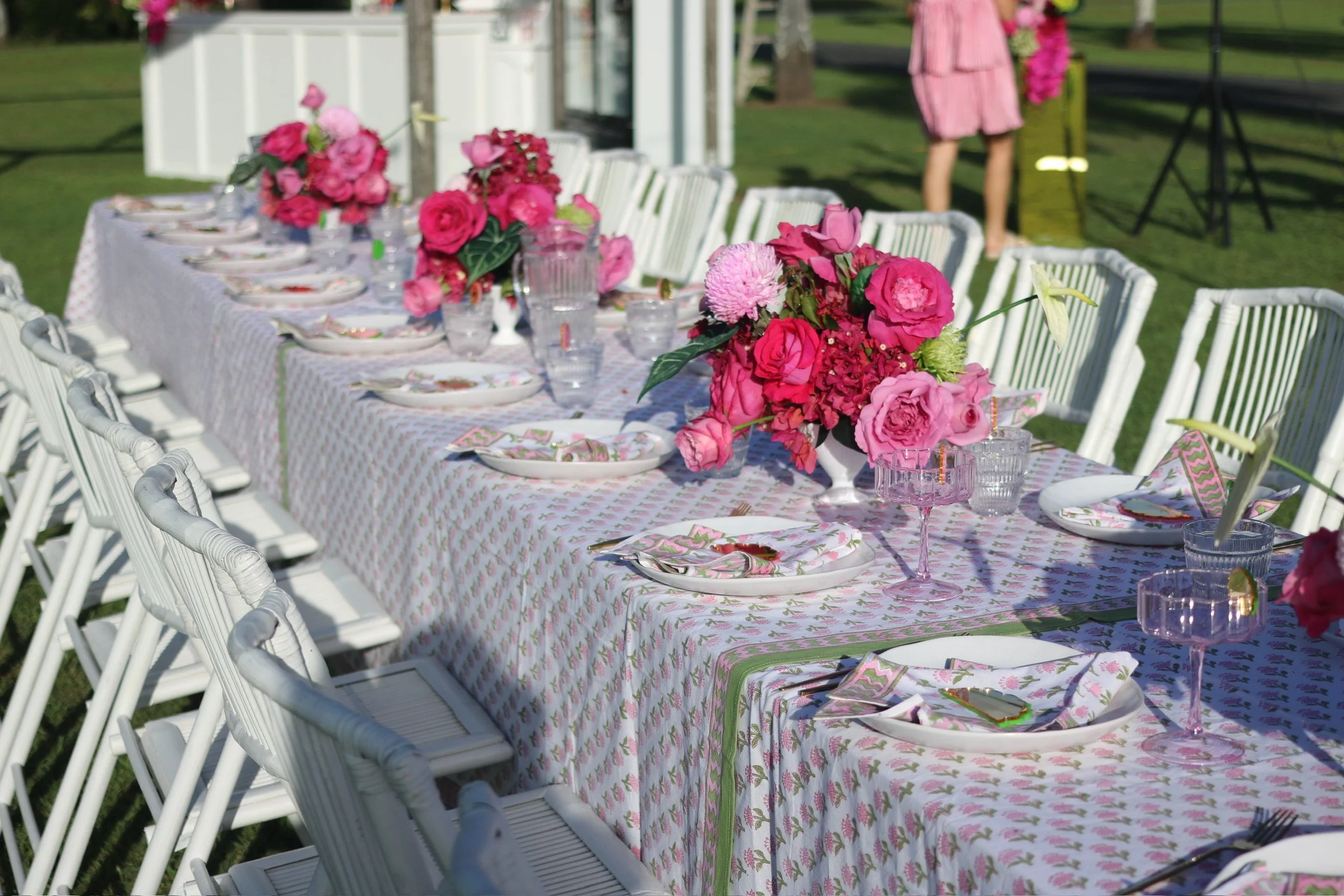 An elegantly set outdoor dining table with a pink floral tablecloth, pink floral centerpieces, and matching napkins, surrounded by white chairs on a grassy area with a person in a pink dress in the background.