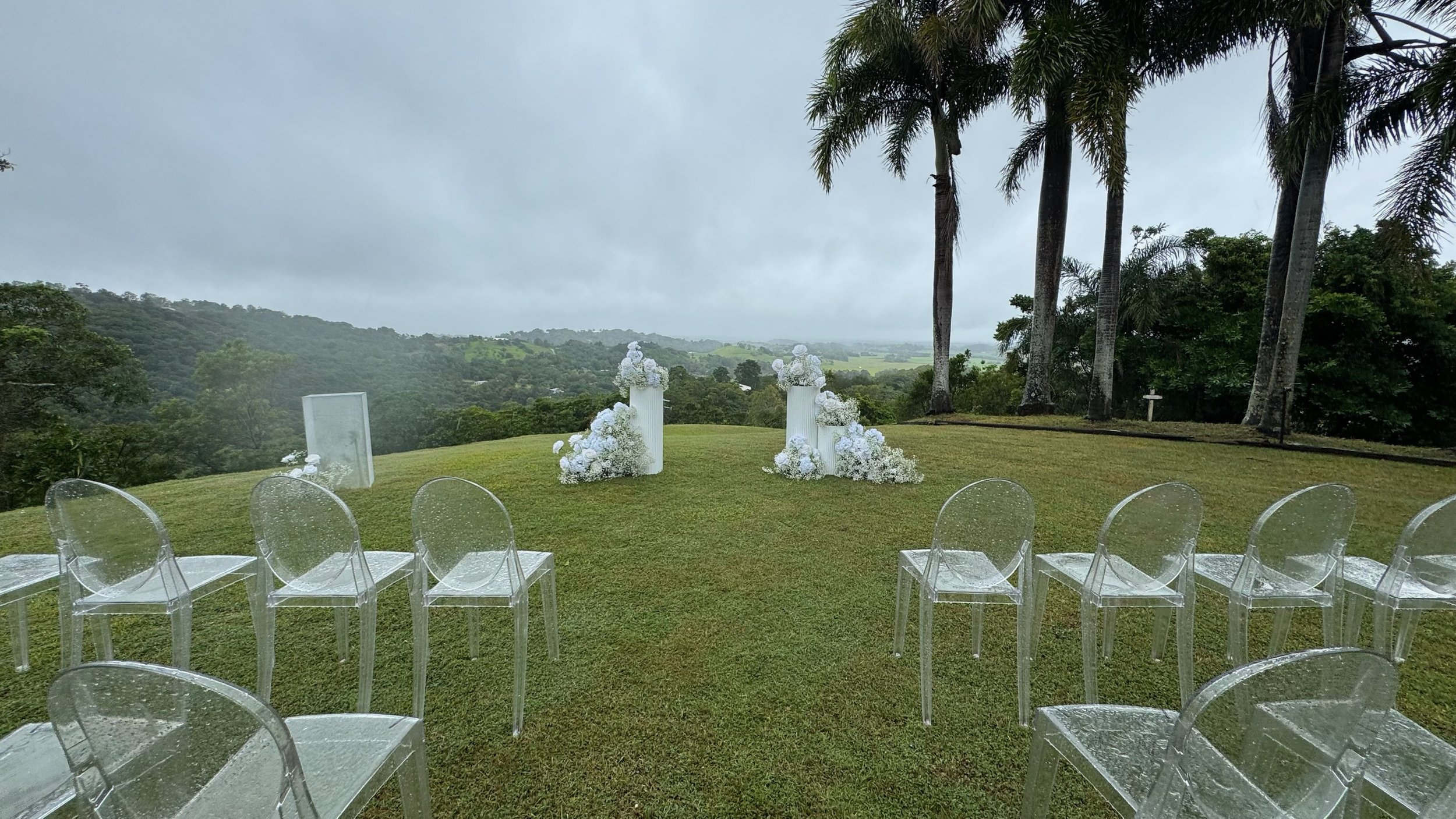 Outdoor wedding ceremony setup with transparent ghost chairs arranged in rows on a grassy hill, floral arrangements on white pedestals, and tall palm trees in the background on a cloudy day.