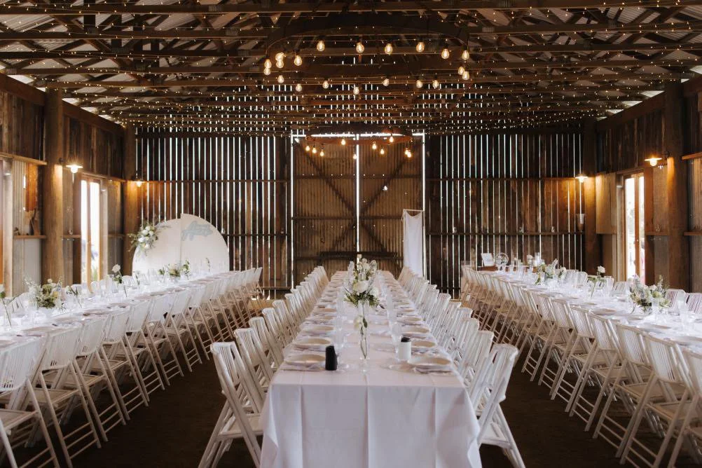 Long banquet tables with white tablecloths, floral centerpieces, and white chairs inside a rustic barn decorated with string lights and Edison bulbs hanging from the ceiling.