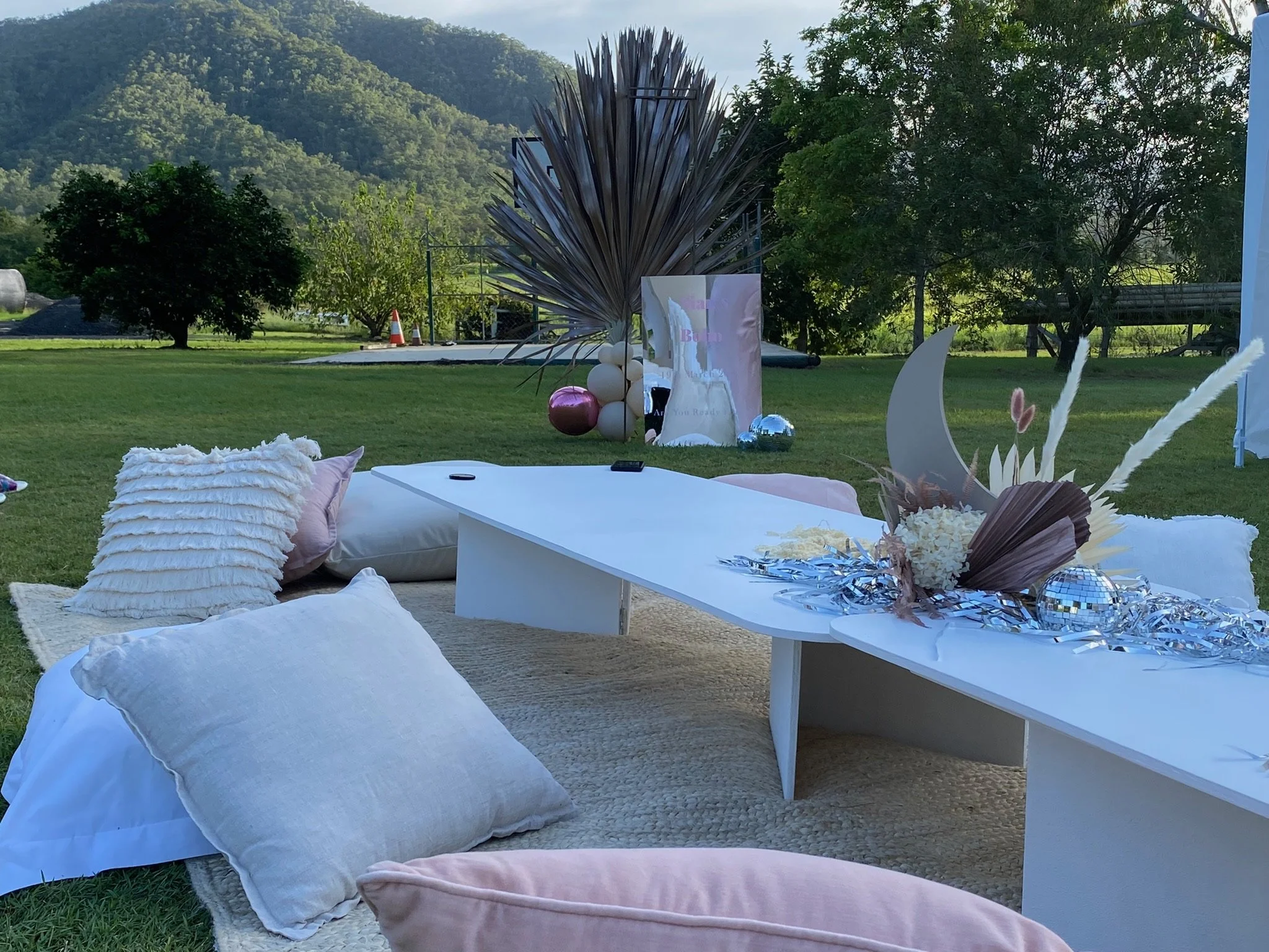 Outdoor event setup with white pillows, a white table decorated with silver streamers, a moon and floral arrangement, and balloons, on grass with trees and mountains in the background.