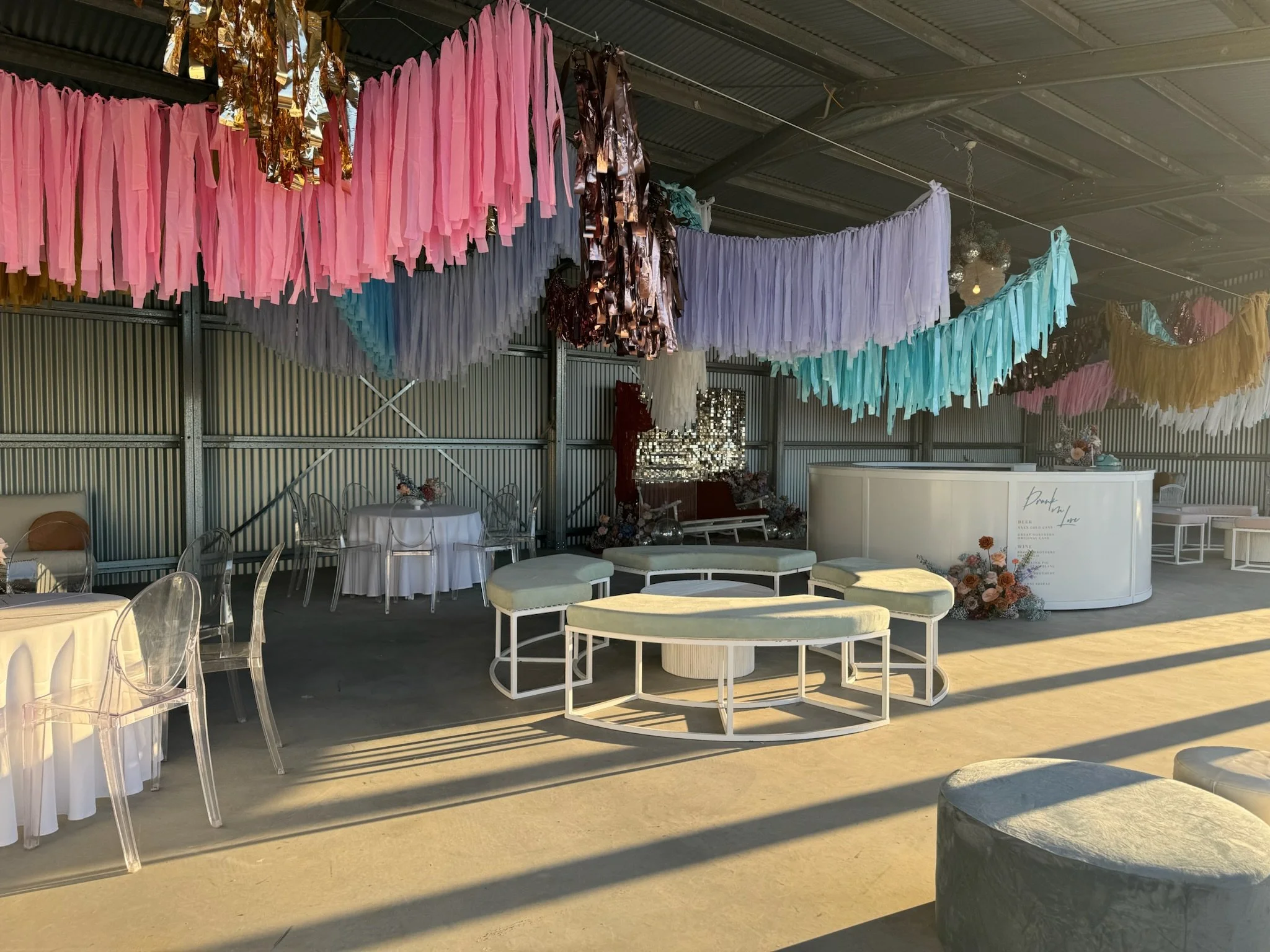Colorful paper tassel decorations hanging inside a decorated venue, with tables and transparent chairs, and a white curved counter with floral arrangements.