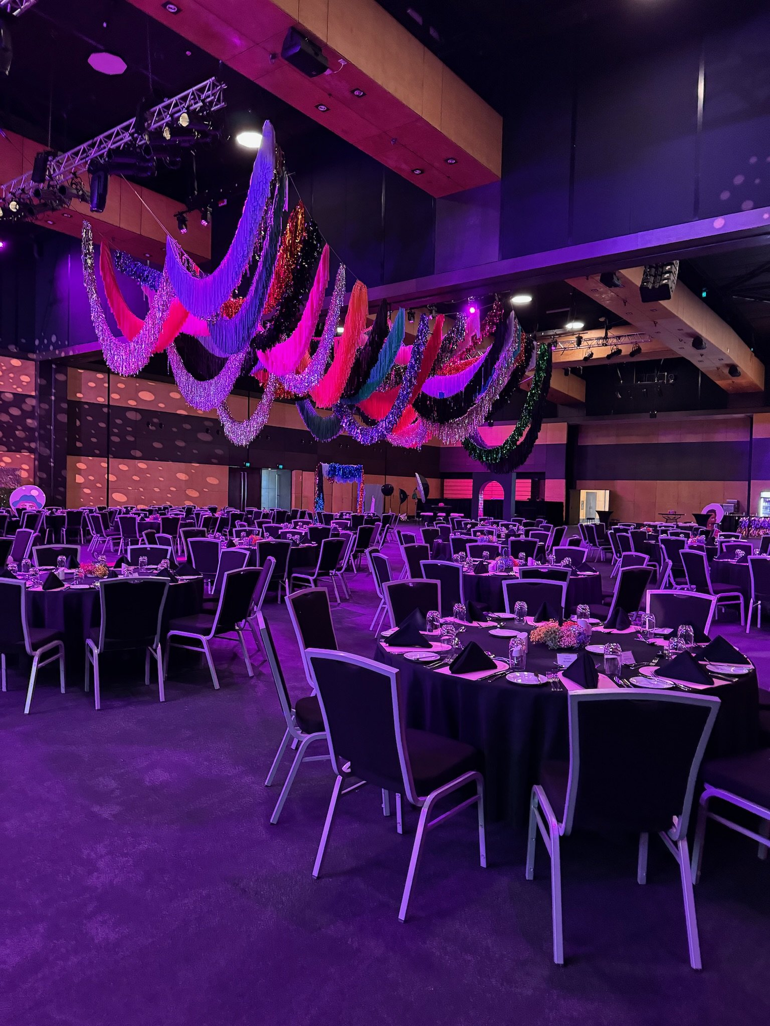 A large banquet hall decorated with black tablecloths, round tables set with plates, glasses, and black napkins, and colorful hanging fabric streamers and lights, indicating a festive event or celebration.