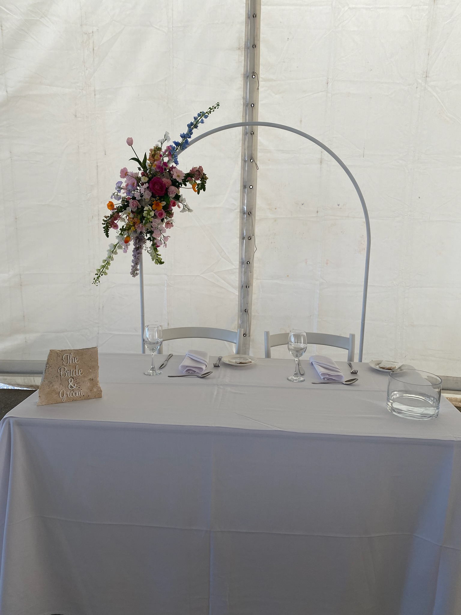 Wedding reception table decorated with a floral arrangement, wine glasses, plates, and napkins, with a sign that says 'The Bride & Groom'.