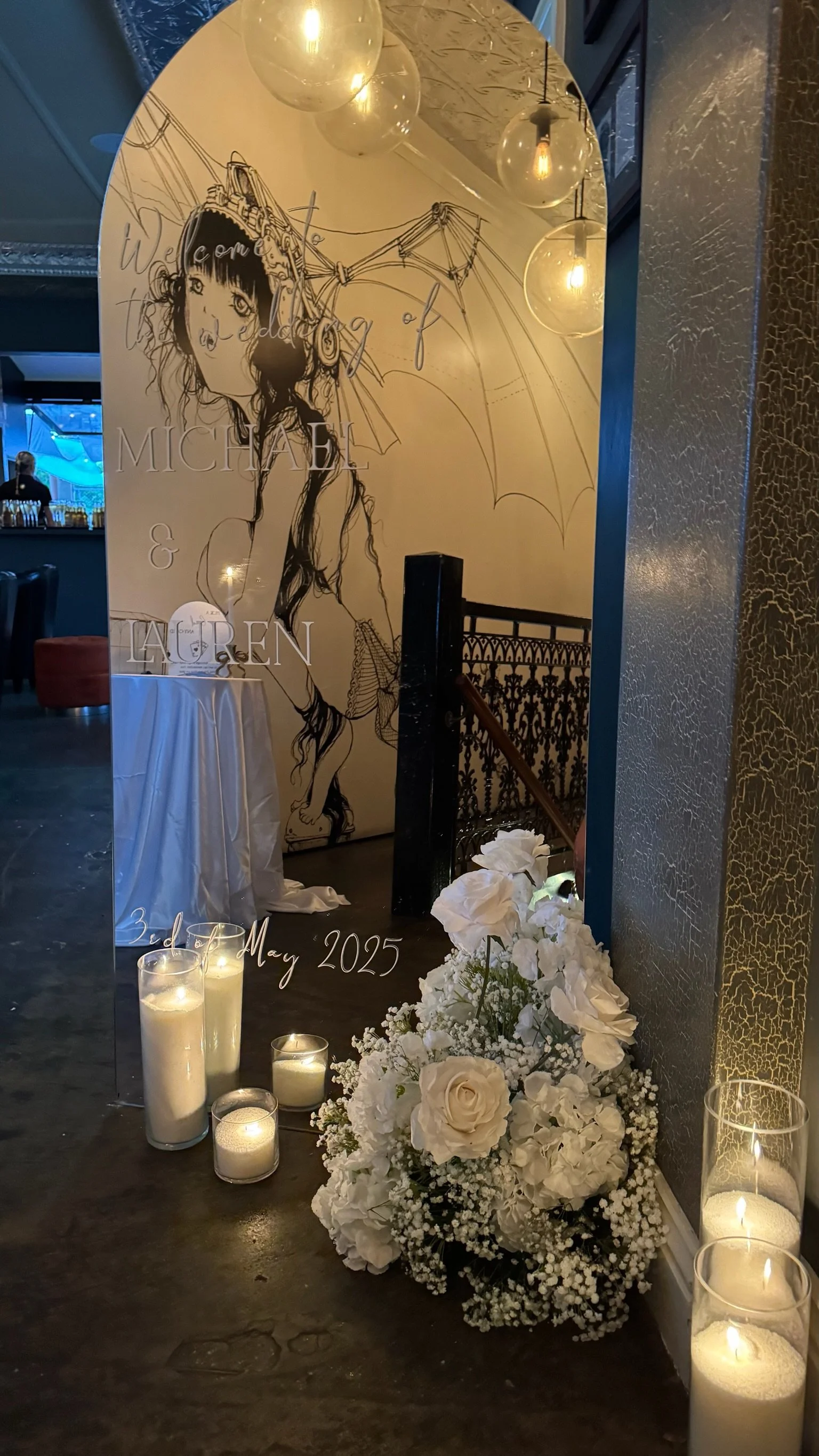 A decorative mirror with a drawing of a girl holding an umbrella, with white flowers and candles in front, and a wedding invitation for Michael and Lauren on May 3rd, 2025.