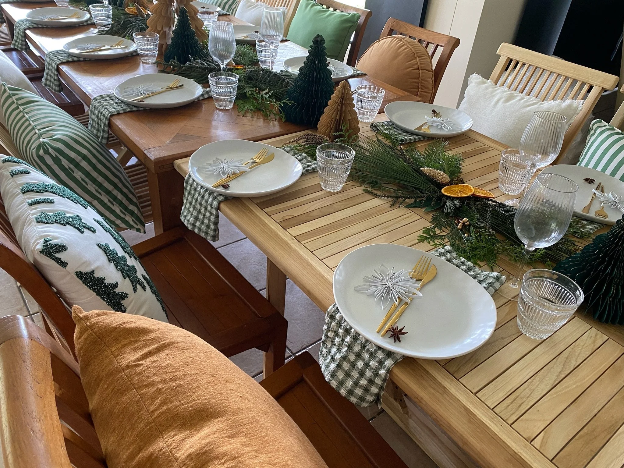 A decorated dining table set for a holiday meal with white plates, gold utensils, glassware, and green checked napkins. The table has a festive centerpiece with greenery, pinecones, dried orange slices, and star anise. Cushions and pillows with holid