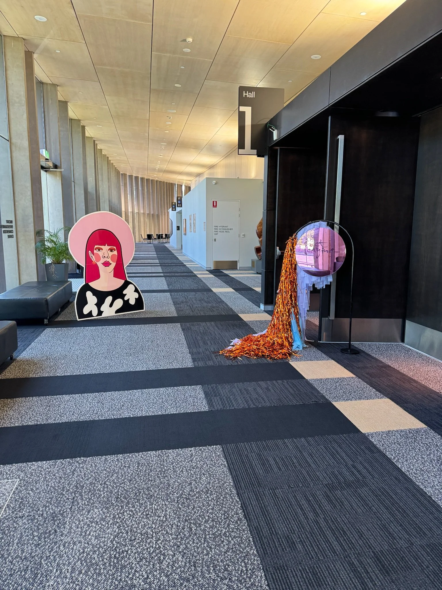 Empty modern hallway in a building with decorative art displays, including a cartoon portrait of a woman with red hair and a large pink hat, and colorful textured sculptures.