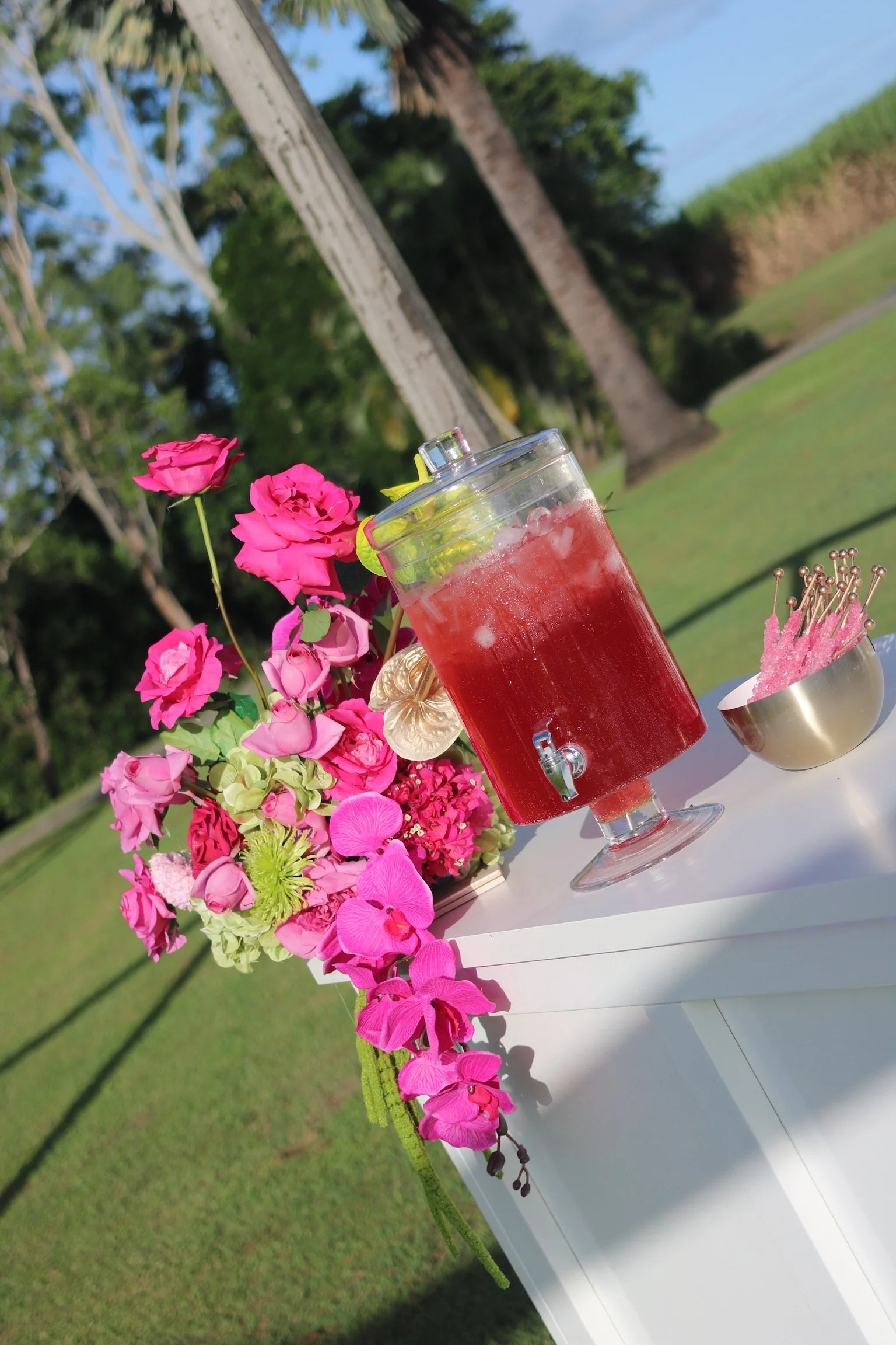 A pink and green floral arrangement, a glass beverage dispenser with pink drink and ice, and a bowl of pink pins are displayed on a white table outdoors with trees and grass in the background.