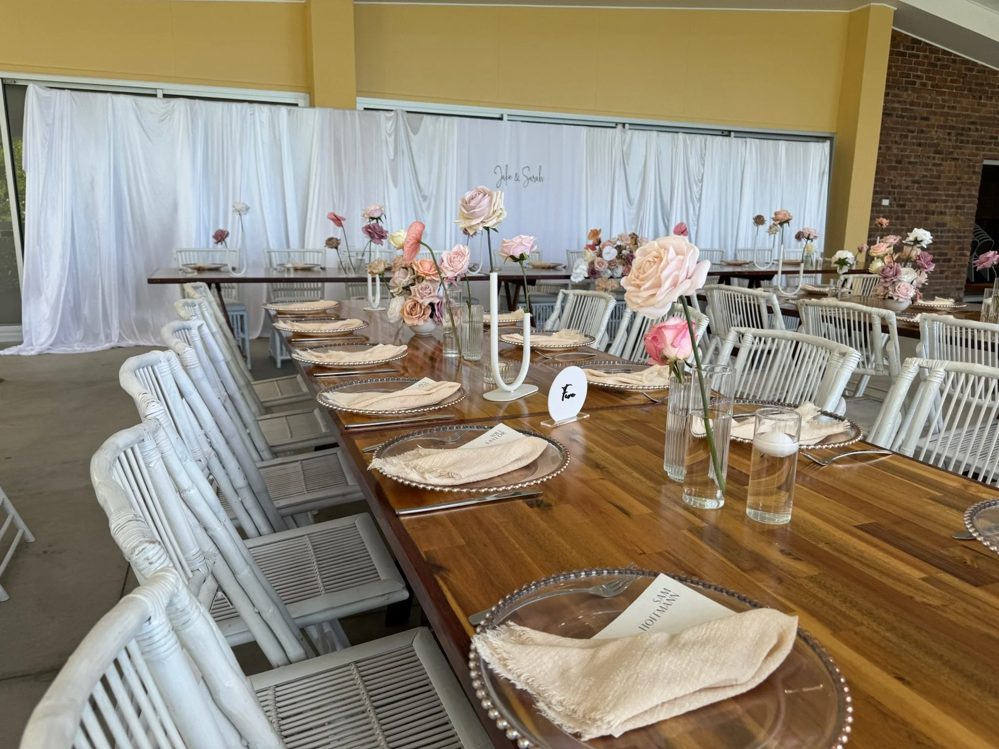 Wedding reception table setup with floral centerpieces, plates, napkins, and place cards in a decorated venue.