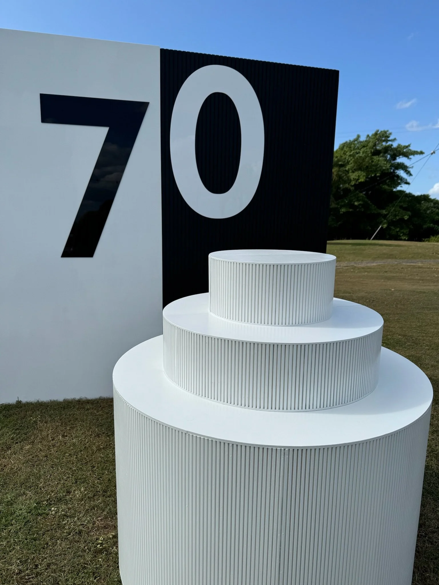A set of three white, ribbed, circular platforms arranged in decreasing height in front of a backdrop with large black and white letters 'Z' and 'O' on a sunny day with a grassy field and blue sky in the background.