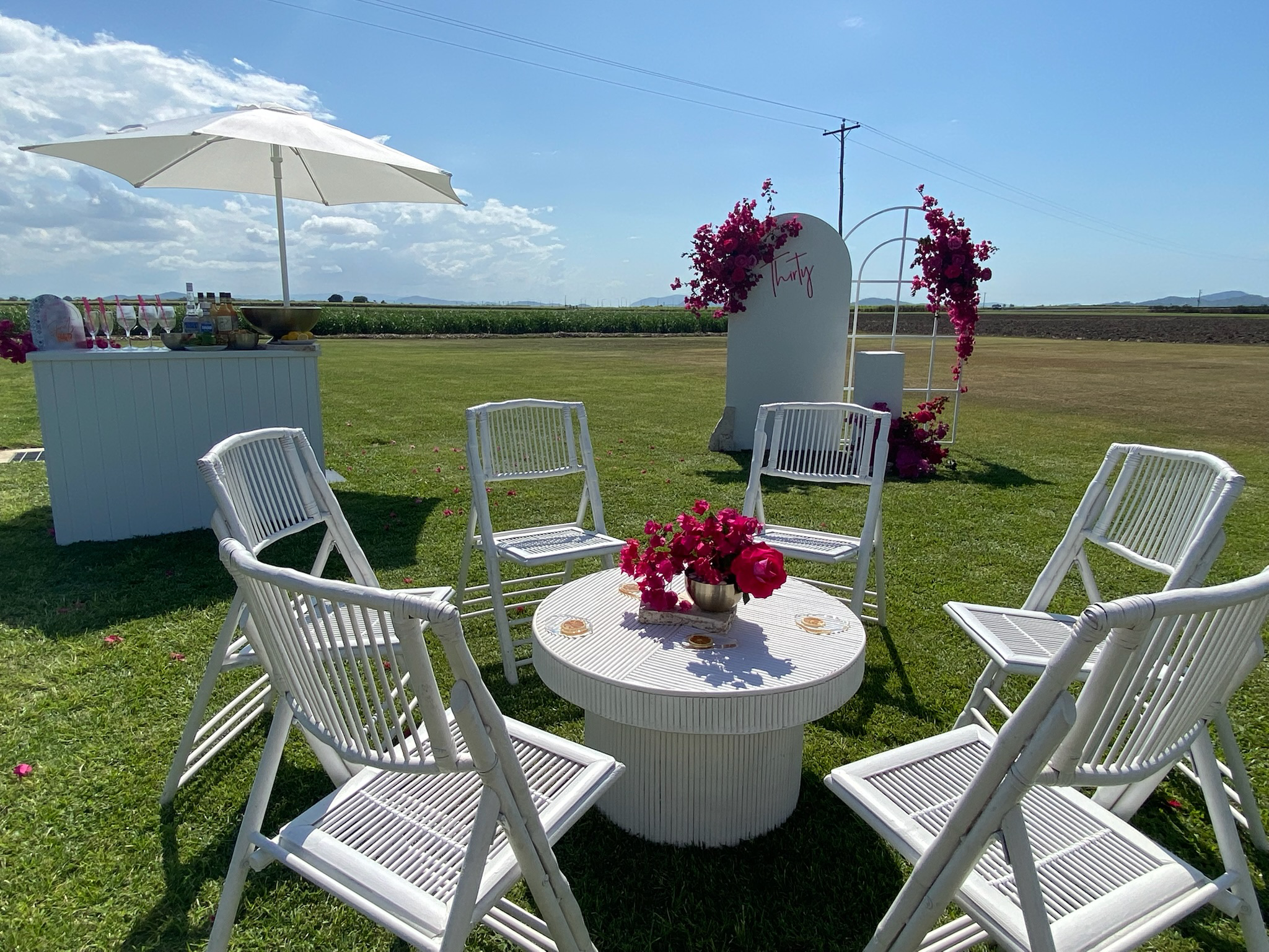 Outdoor setup for a celebration with white chairs surrounding a round table with pink flowers, a white umbrella, and a refreshment station, on a green grassy field with a decorative backdrop and open sky