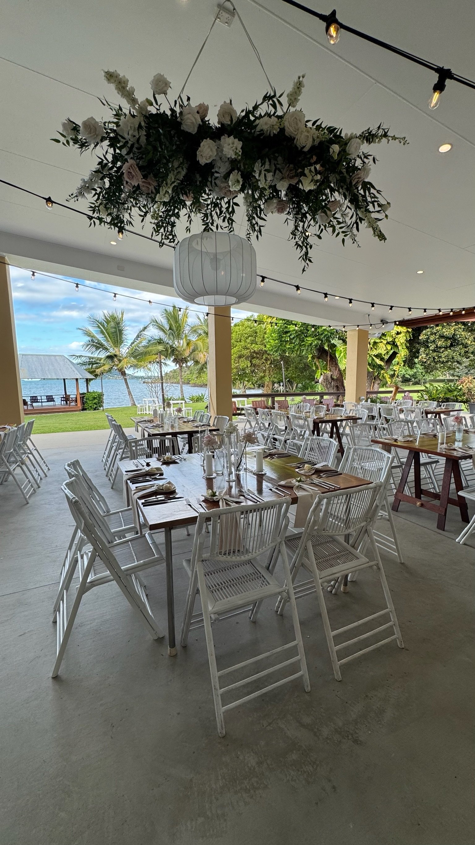 Outdoor wedding reception setup with tables, white chairs, and floral decorations by a waterfront.