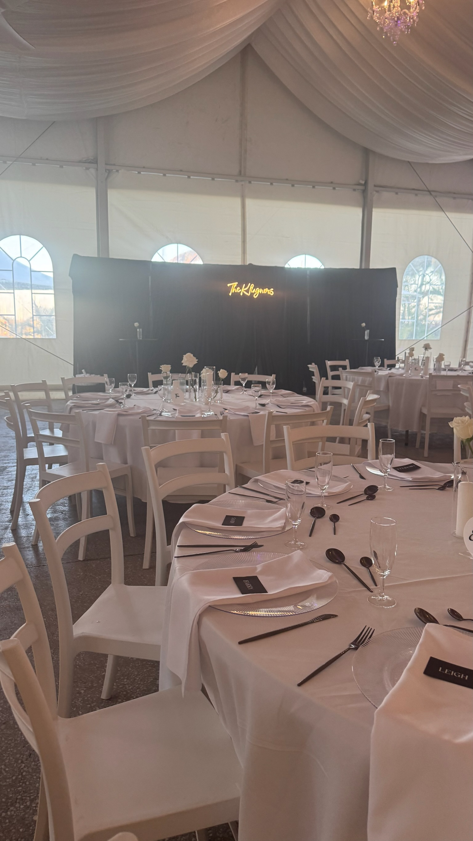 Wedding reception area inside a large tent with decorated tables, white chairs, and a stage with a sign that reads 'The Klagners.'