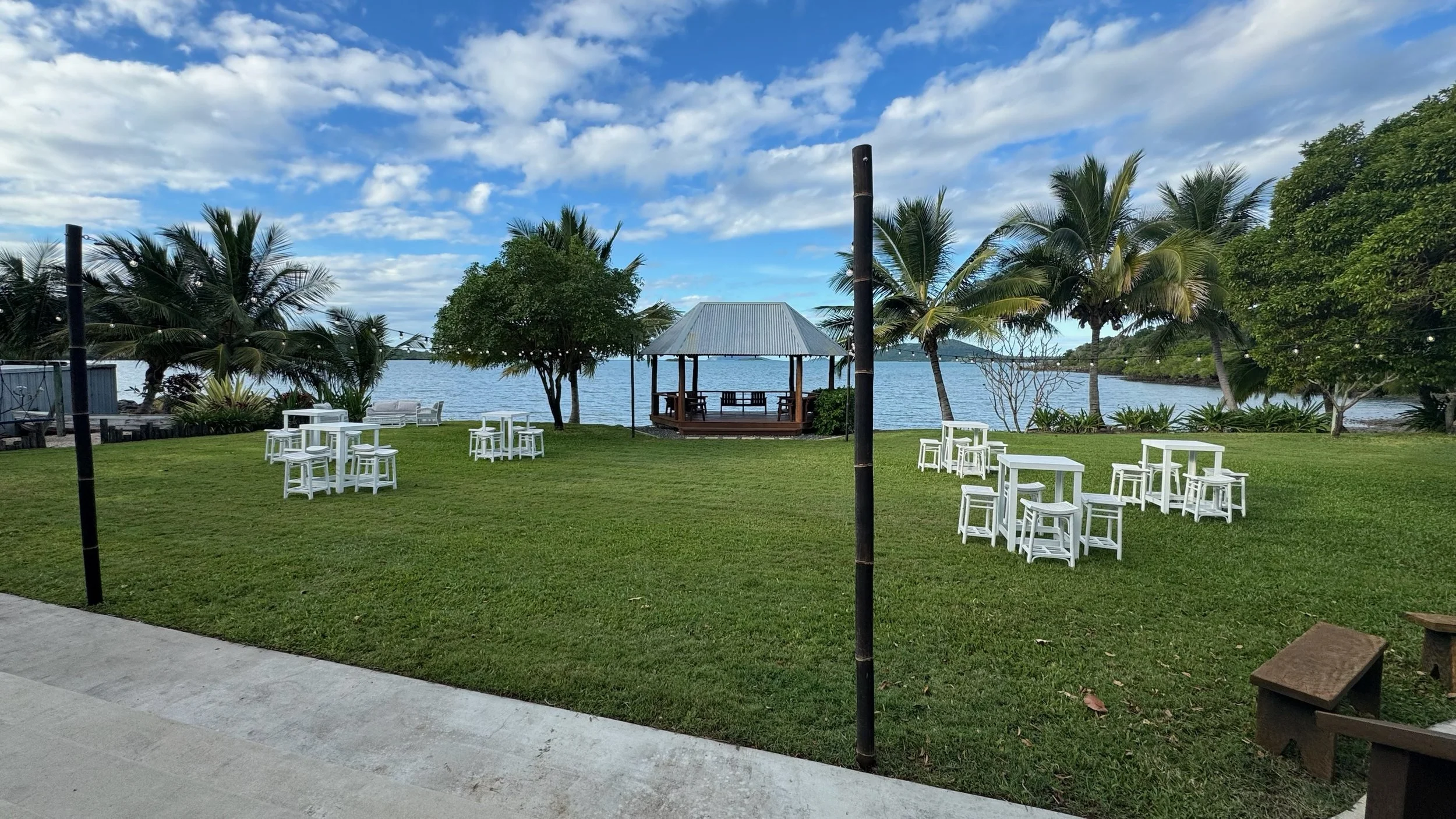 Lawn area with white tables and chairs near the water, and a wooden gazebo with a metal roof in the background, surrounded by palm trees and other lush vegetation, under a partly cloudy sky.
