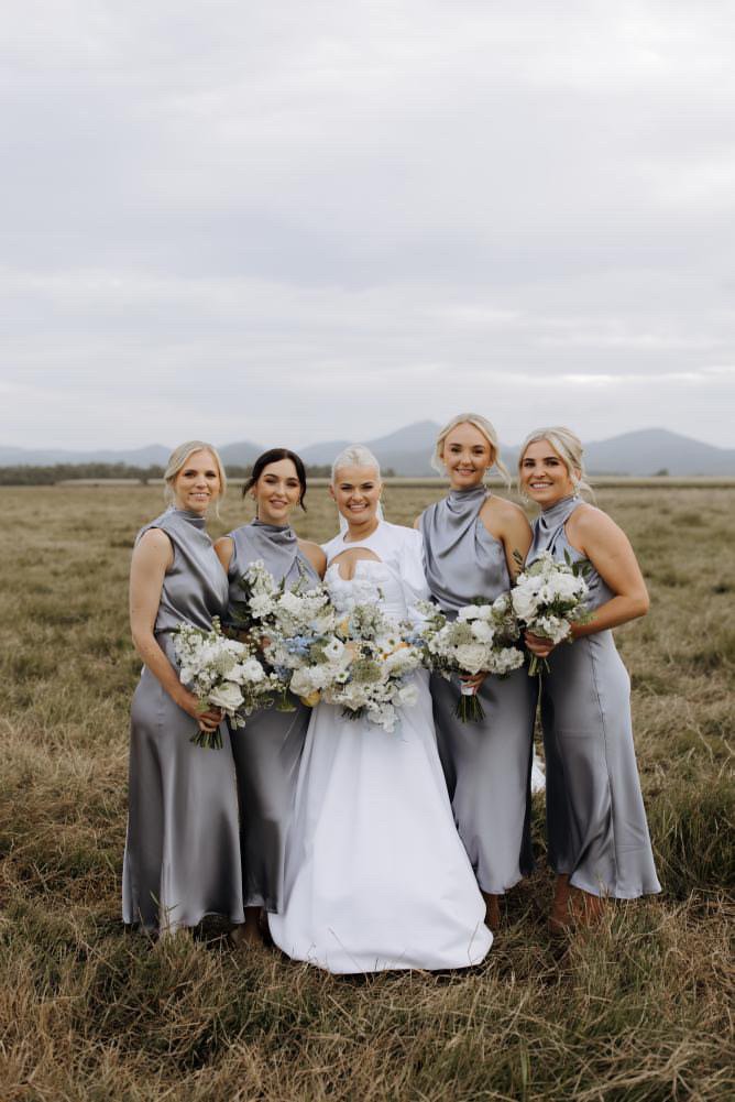 A bride and four bridesmaids standing together outdoors on a grassy field with mountains in the background, each holding a bouquet of white flowers. The bride is in a white gown, and the bridesmaids are in matching gray satin dresses.