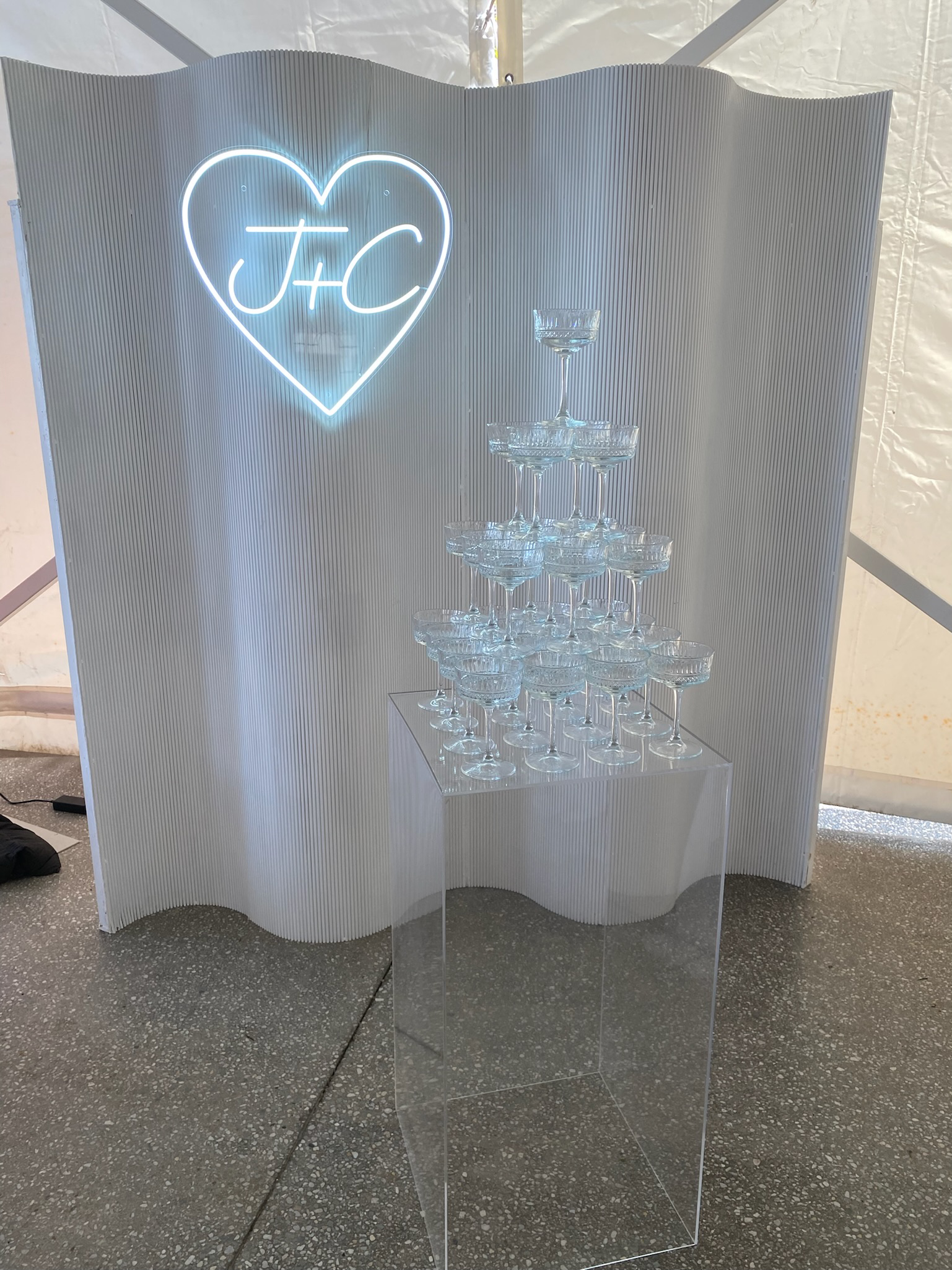A display of champagne glasses arranged in a pyramid shape on a transparent stand. Behind it is a textured white backdrop with a neon sign in the shape of a heart containing the initials 'J+C'.