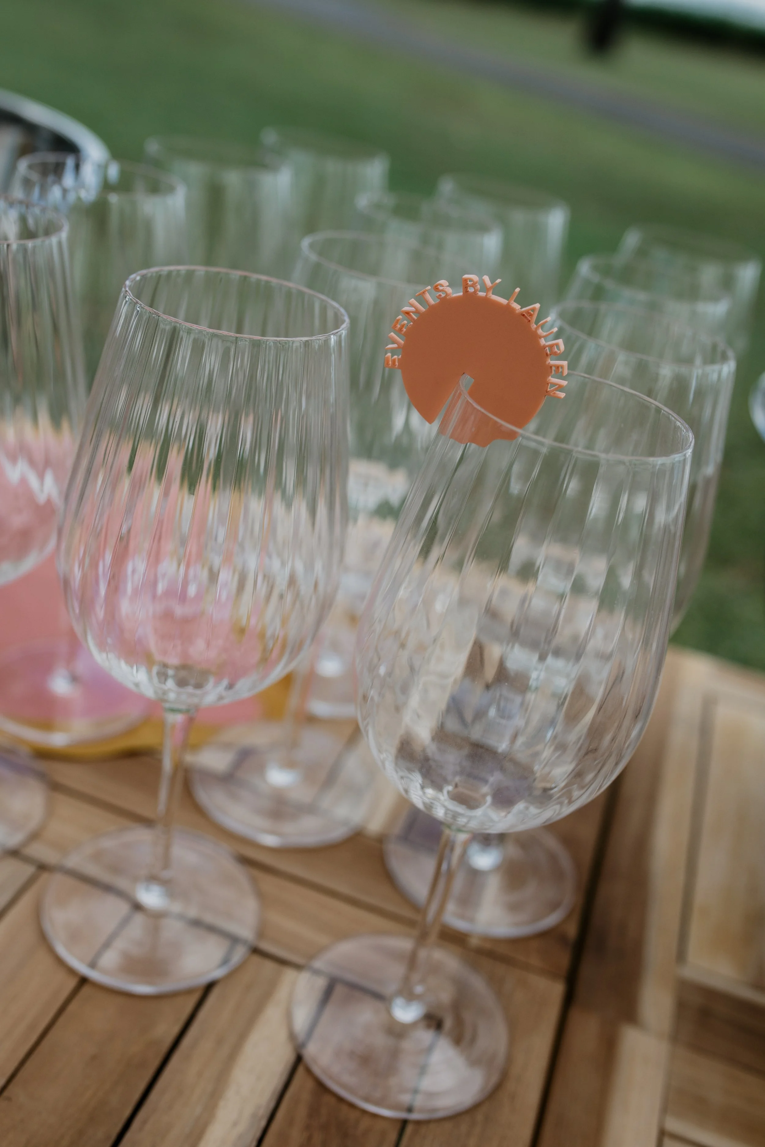 Multiple empty champagne flutes on a wooden table at an outdoor event, with a small sign on one glass that reads 'Events by Lauren'.