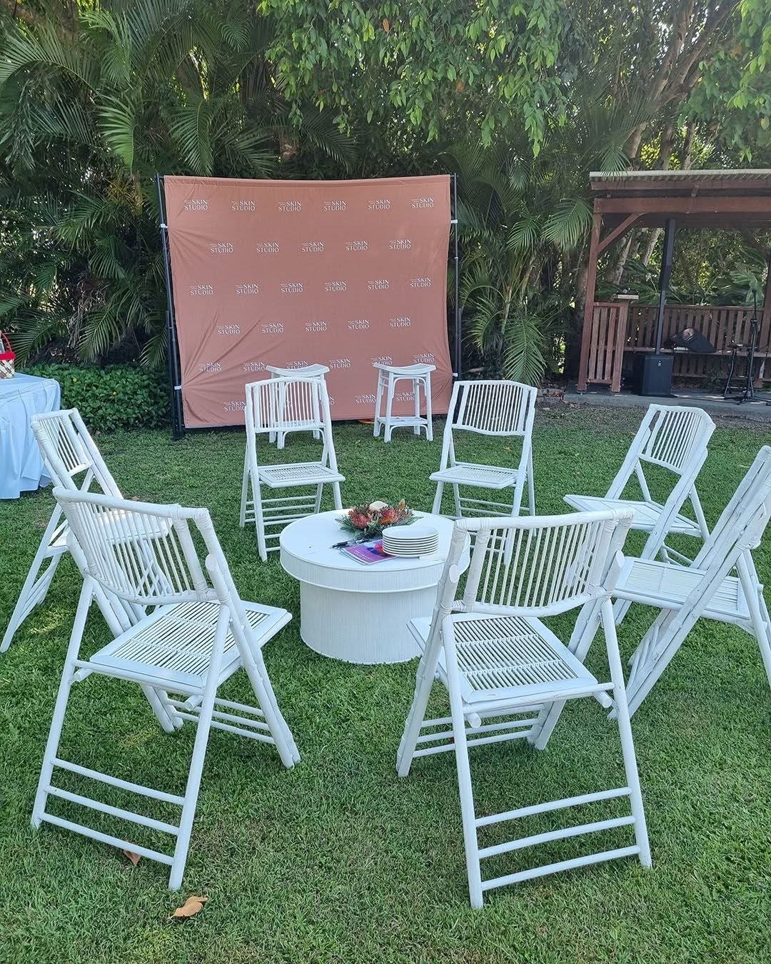 Outdoor event setup with white chairs arranged in a circle around a white round table with plates and a floral centerpiece. A large brick backdrop with the words 'Skin Studio' repeats in the background. There are trees and greenery surrounding the ar