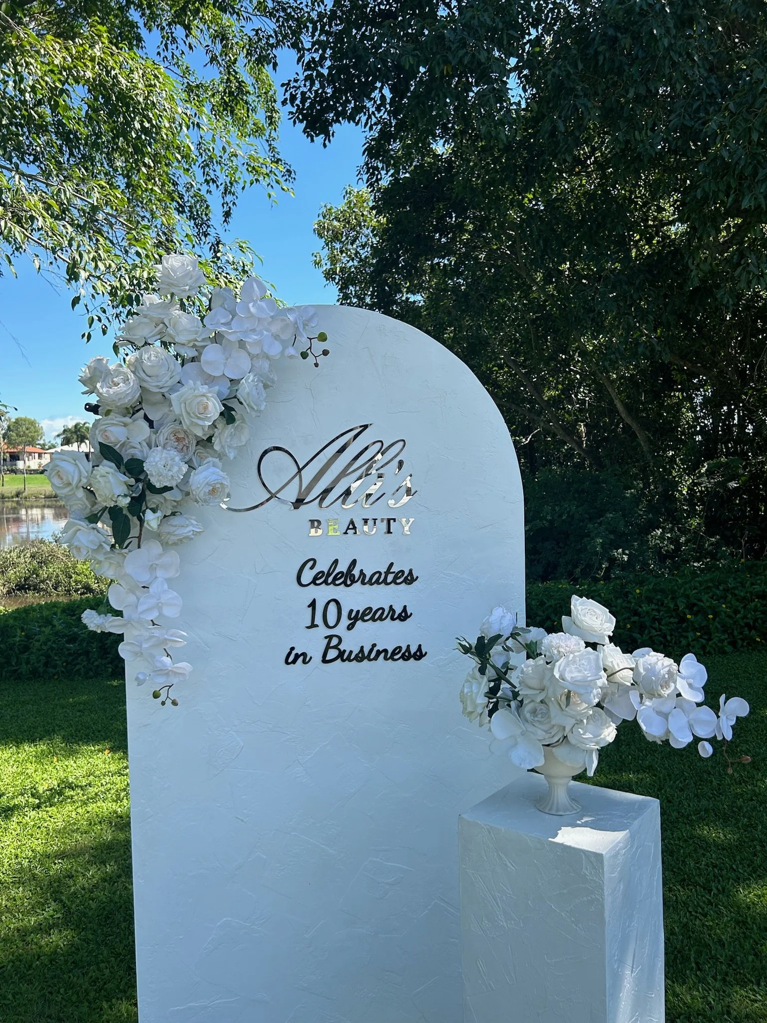 A tall white sign decorated with white flowers and greenery, celebrating 10 years in business for Alik's Beauty, outdoors with trees and a river in the background.