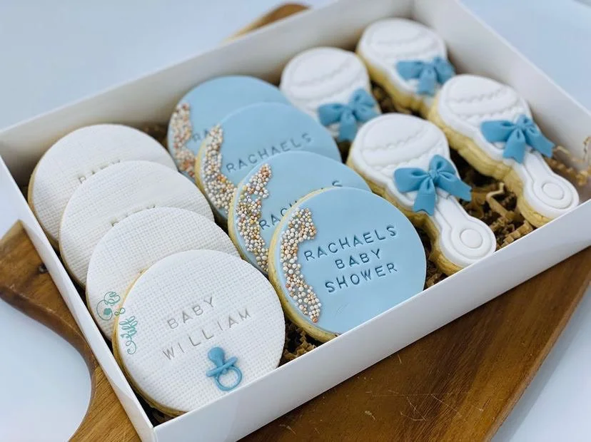 Box of decorated cookies for a baby shower, with designs including baby hats, pacifiers, and onesies in blue and white colors.