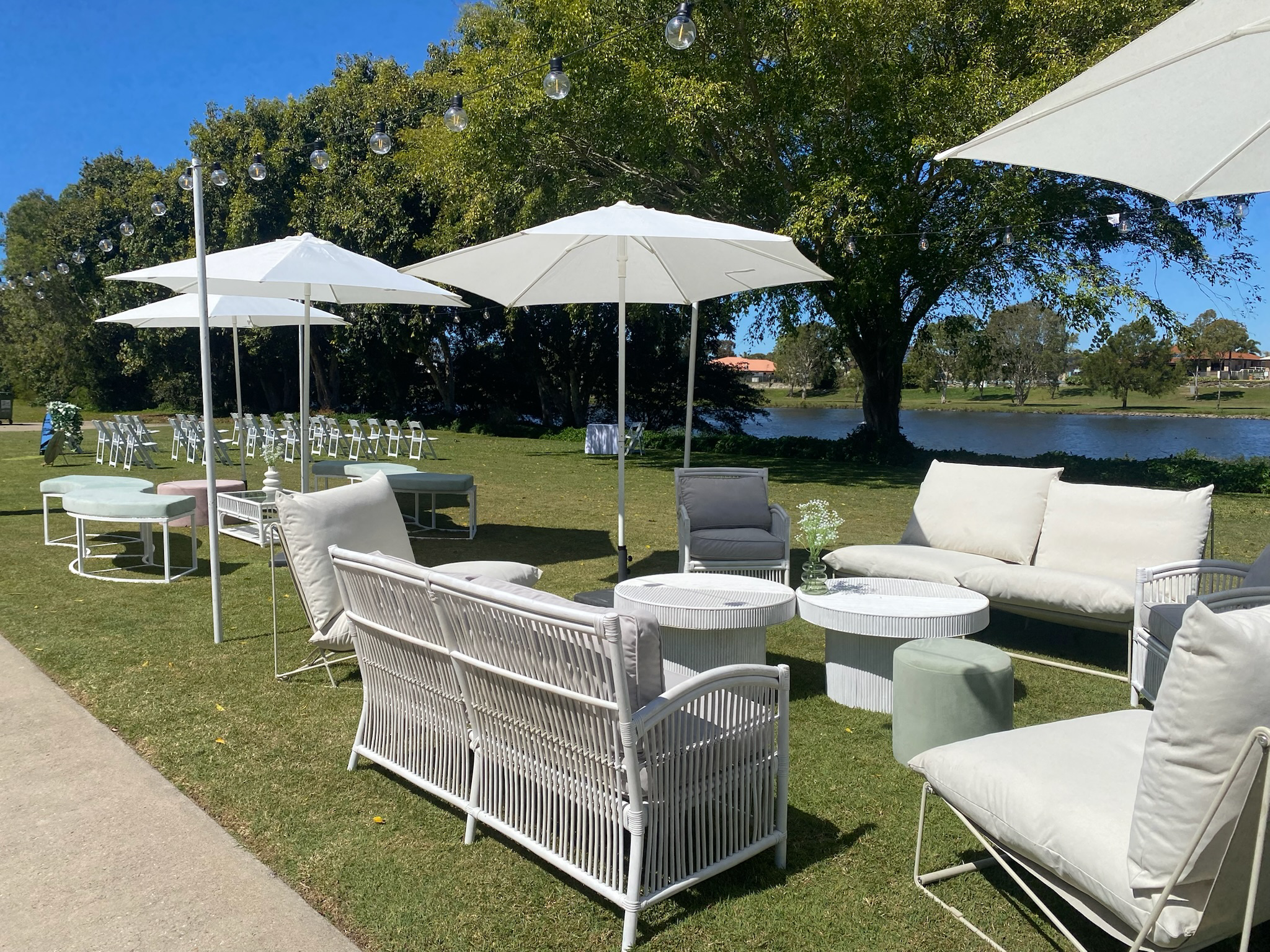 Outdoor seating area with white upholstered chairs, sofas, coffee tables, white umbrellas, and string lights by a lake under a tree on a sunny day.