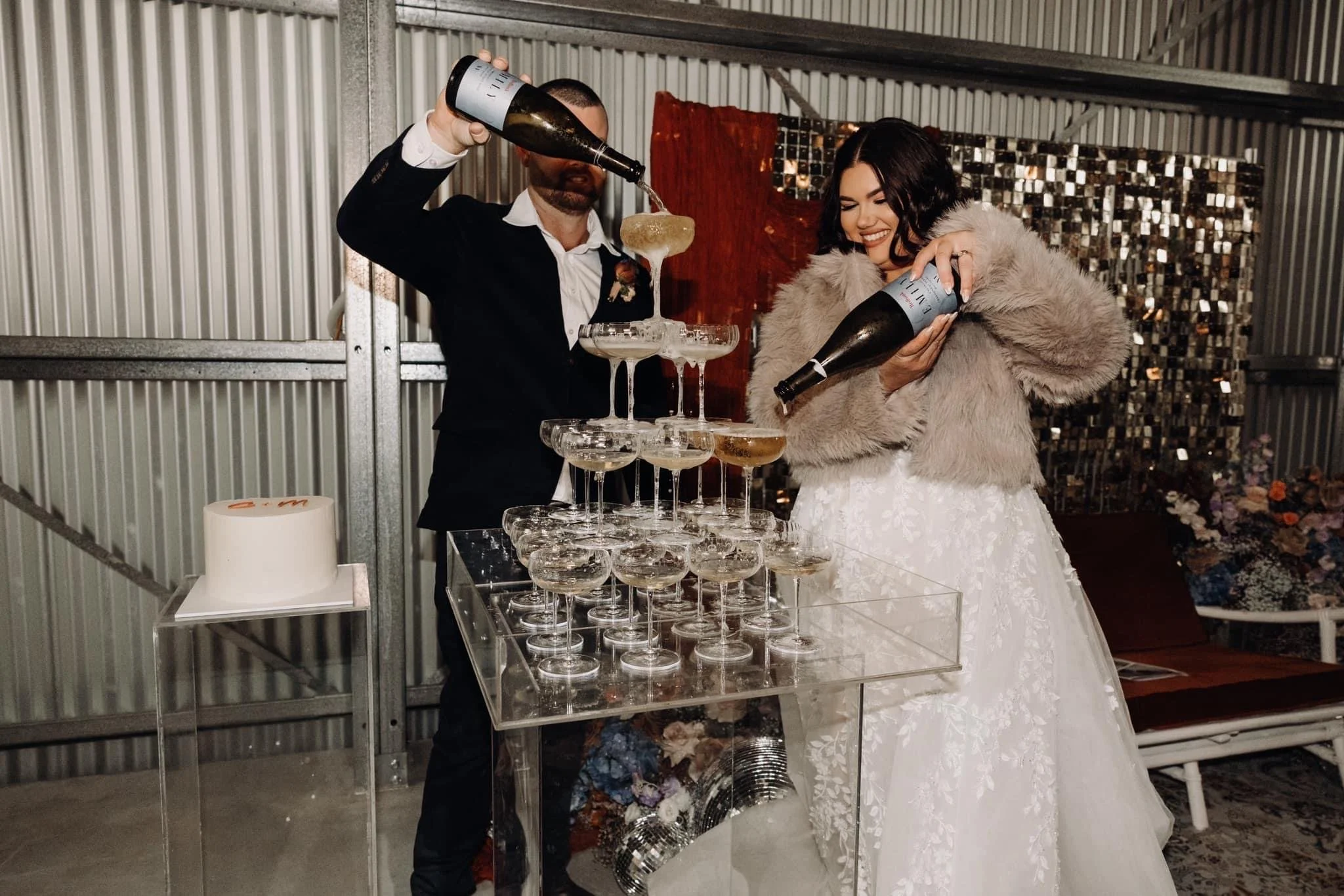 Couple in wedding attire celebrating at a champagne tower during their wedding reception, with a wedding cake nearby.