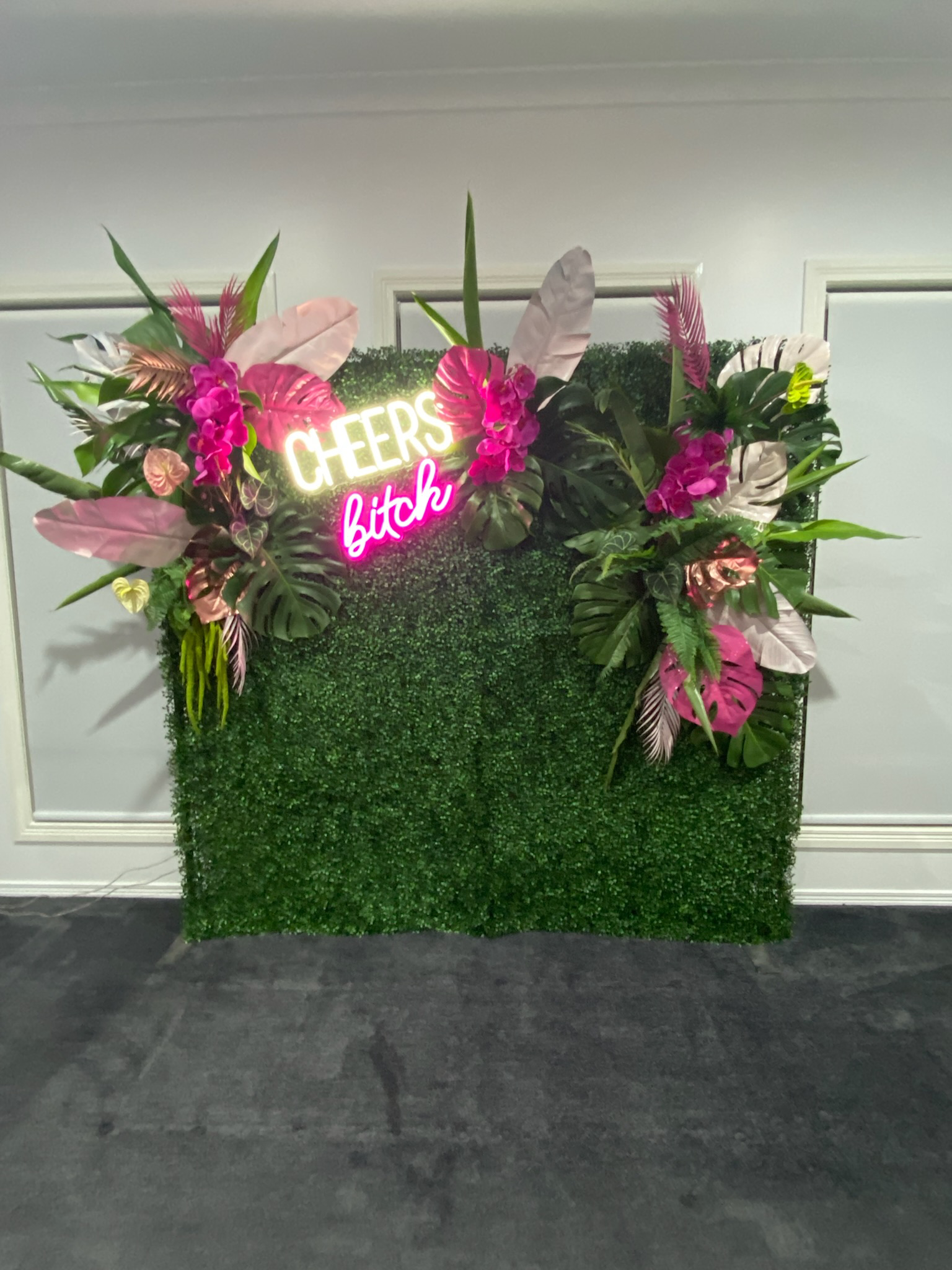 Decorative green hedge backdrop with pink and white leaves, pink flowers, and a neon sign reading 'Cheers bitch' in white and pink.