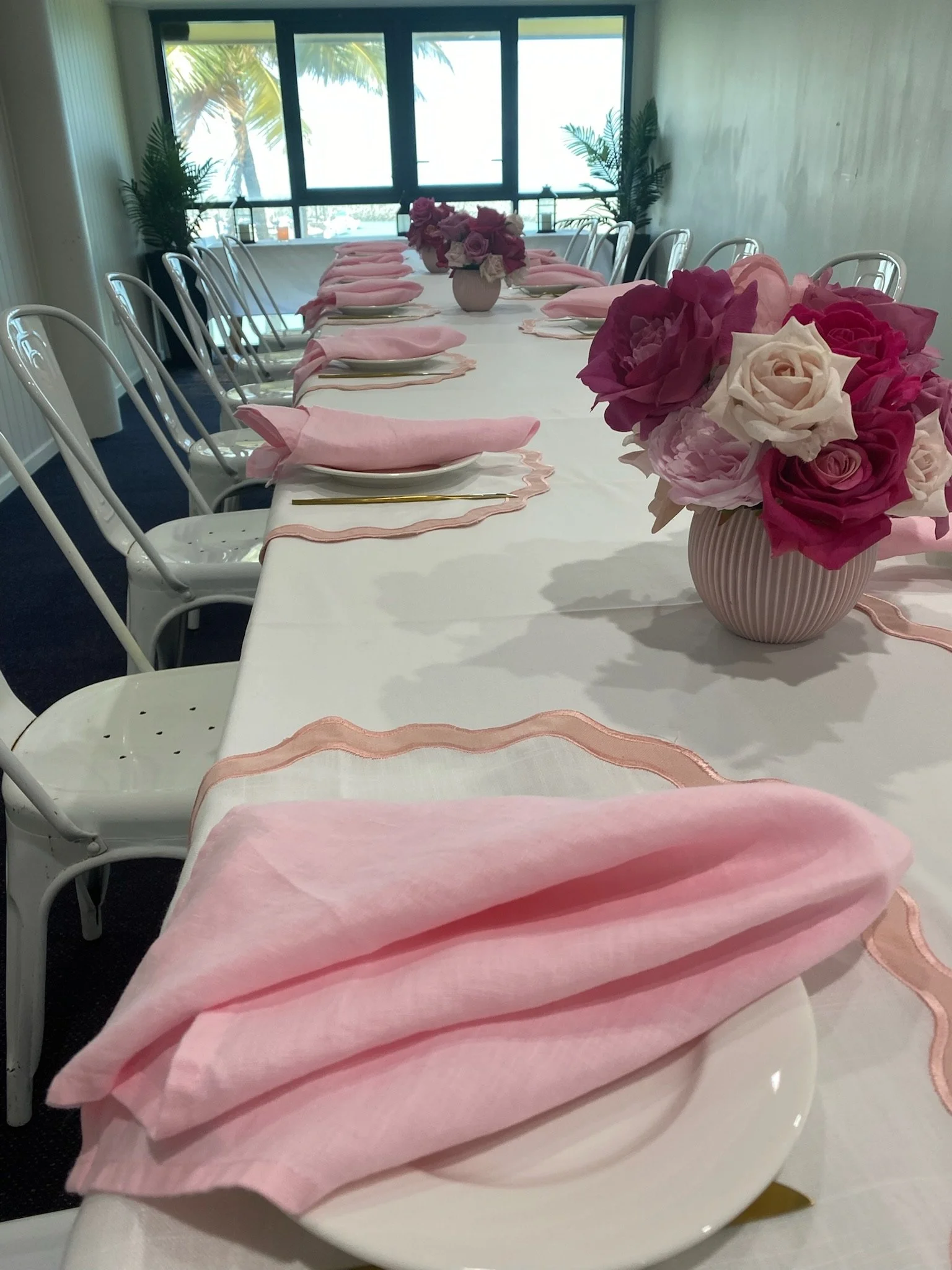 A long dining table decorated for a celebration with pink napkins, gold cutlery, and pink and white flower centerpieces in vases, set near large windows overlooking palm trees.