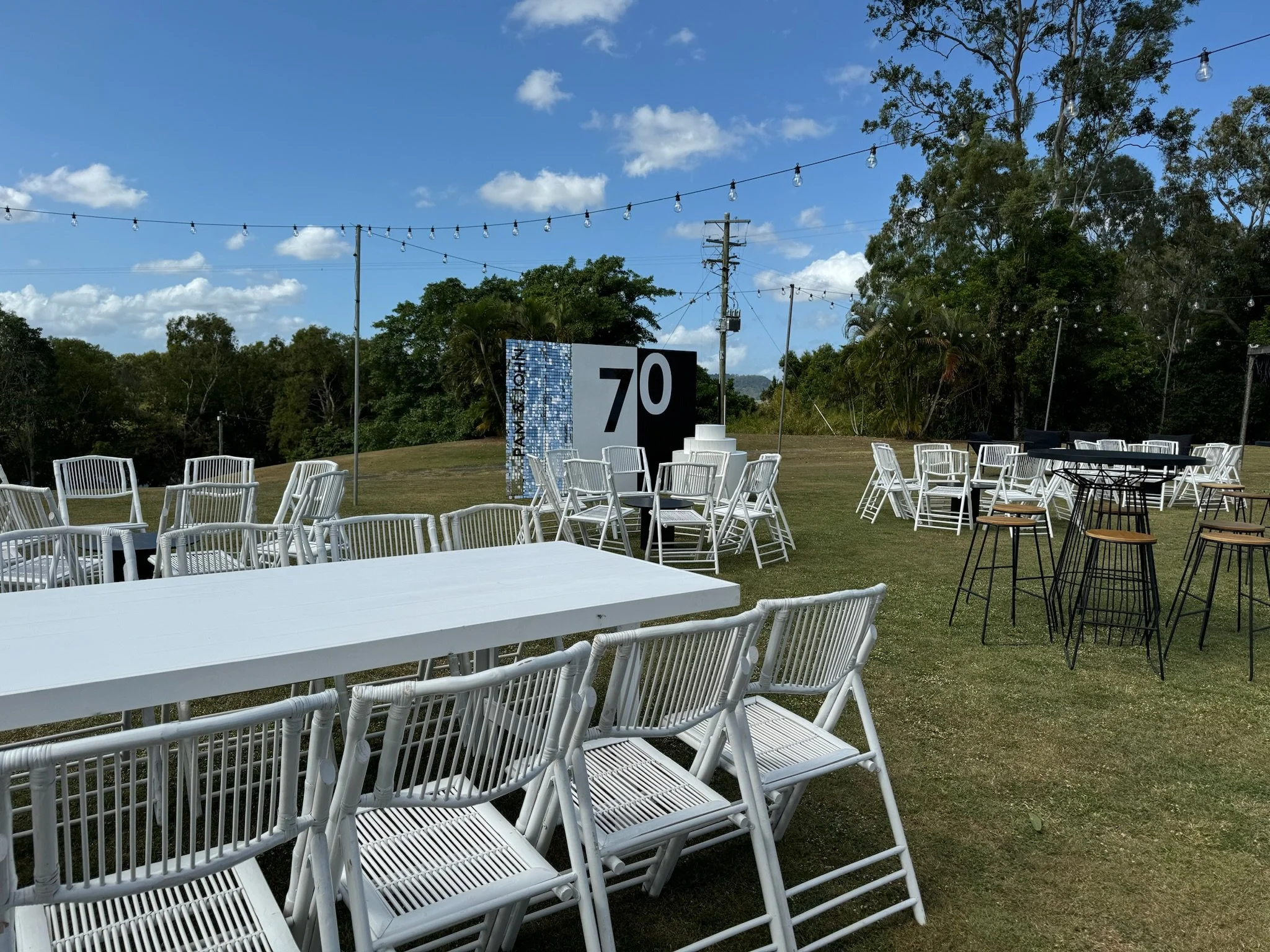 An outdoor event setup on a lawn with white chairs and tables, string lights overhead, a large sign with the number 70, and a backdrop with 