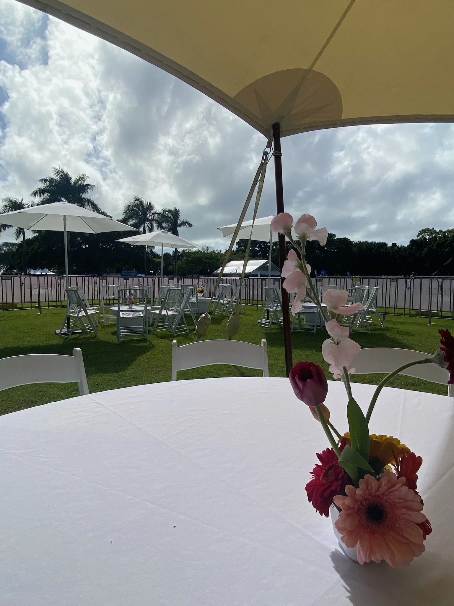 Outdoor event setup with white tables and chairs, large umbrellas, and a flower centerpiece on a white tablecloth, with palm trees and a cloudy sky in the background.