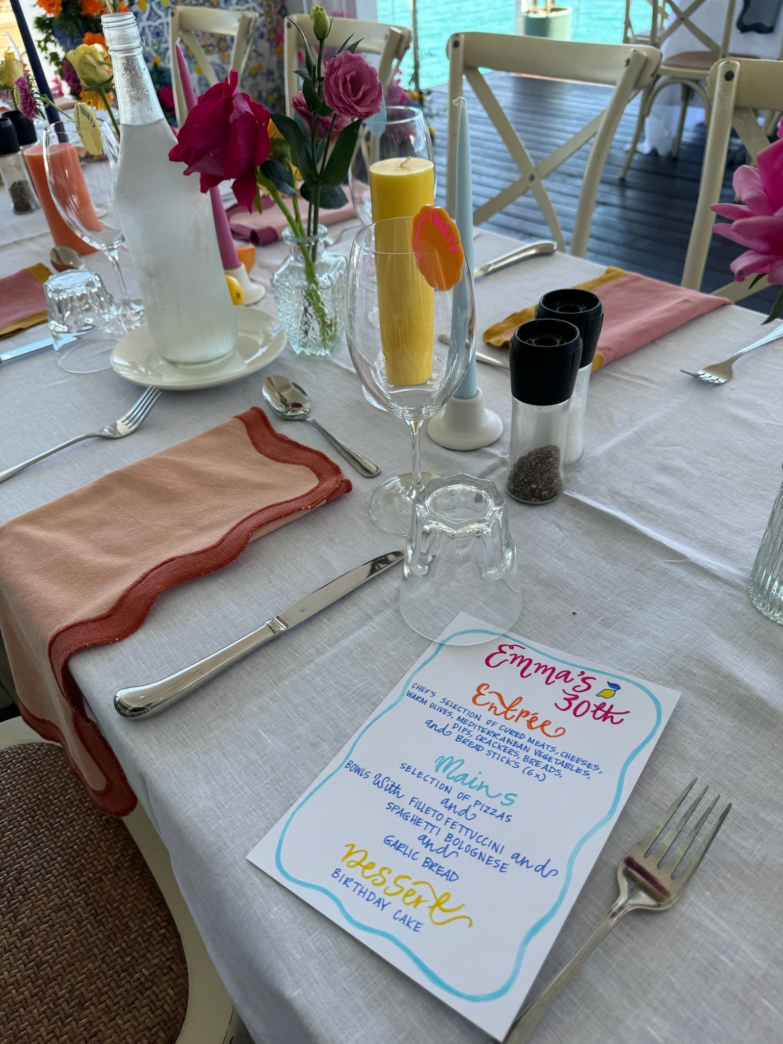 A decorated dining table set with a menu, wine glasses, candles, flowers, and tableware for a celebration, with a view of a lake or pool in the background.