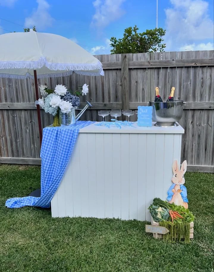 A backyard party setup with a white bar or table against a wooden fence, topped with a blue and white checkered cloth, a large white umbrella, a flower arrangement, champagne bottles in an ice bucket, and glasses, with a decorative bunny figure and a
