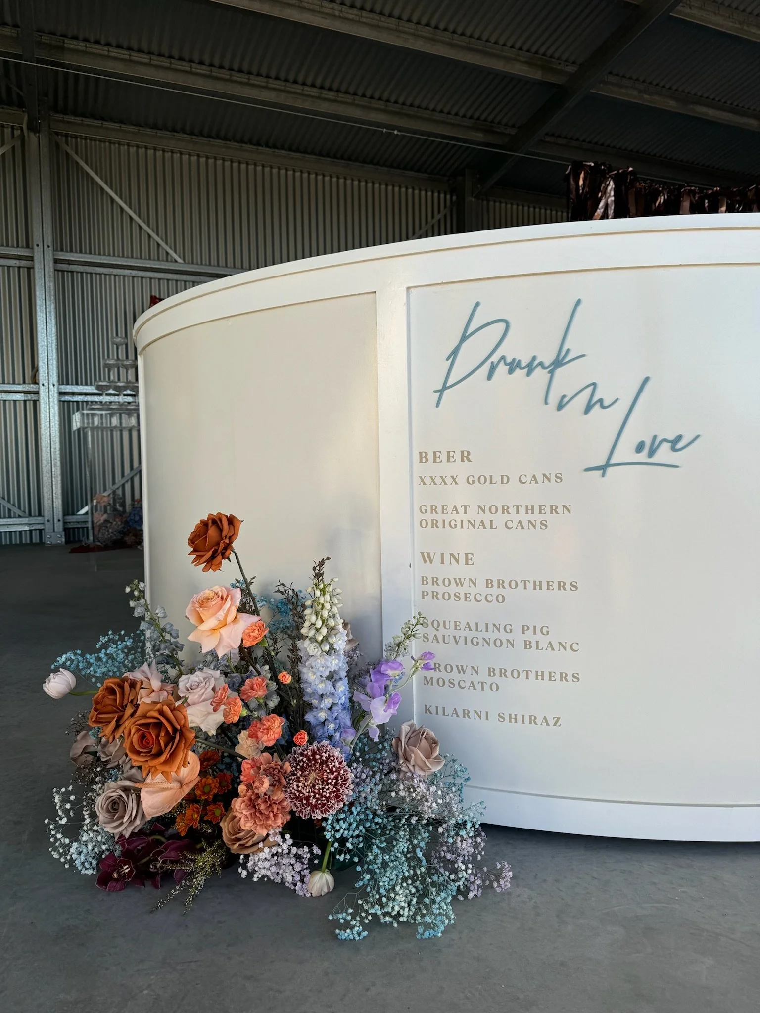 A large floral arrangement with roses, carnations, and baby's breath in shades of pink, orange, purple, white, and burgundy, placed in front of a white structure with a menu titled 'Drink and Love' listing beers and wines. The setup appears in an ind