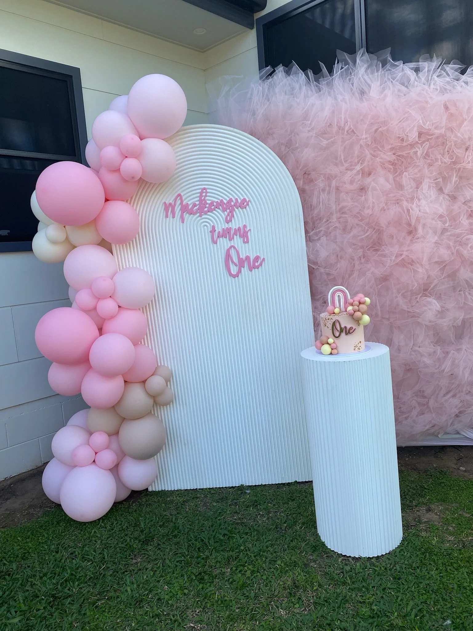 A pink and white balloon arrangement next to a pink birthday cake on a white pedestal, with a backdrop of pink feathers and a white arched display with pink words that say 'Making turns One.'