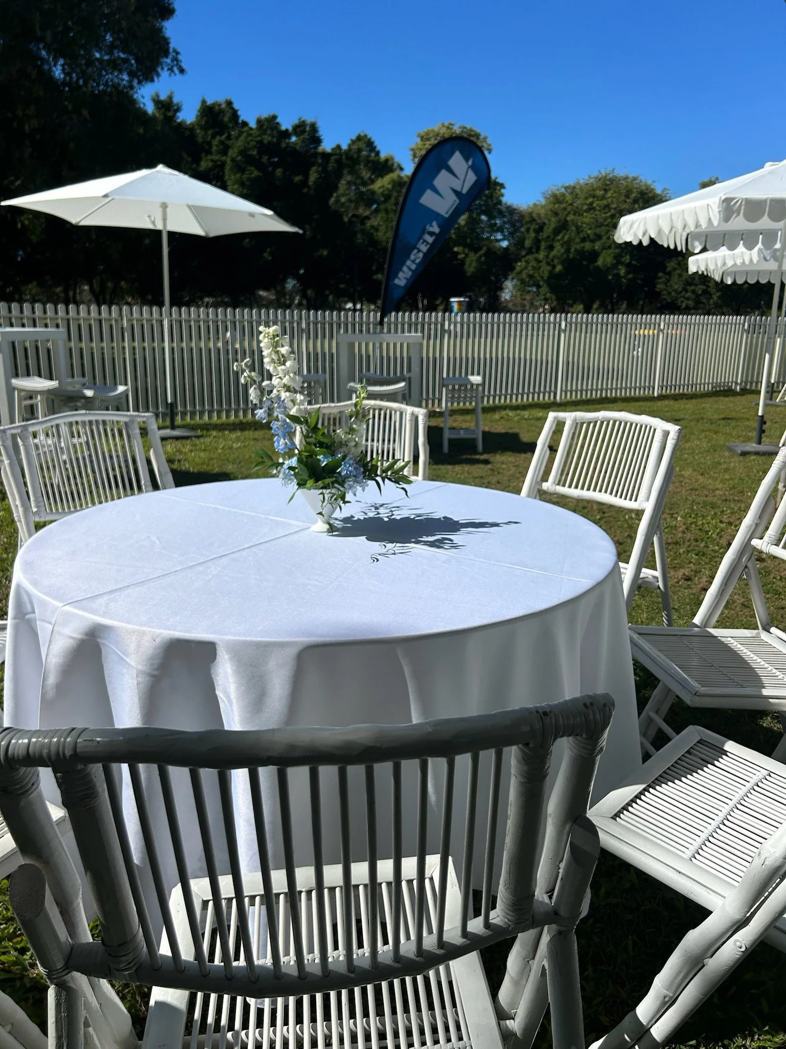 Outdoor event setup with a round table covered in a white tablecloth, surrounded by white chairs, with a floral centerpiece. In the background, there are white umbrellas, a fence, and a blue flag with white lettering, under a clear blue sky.