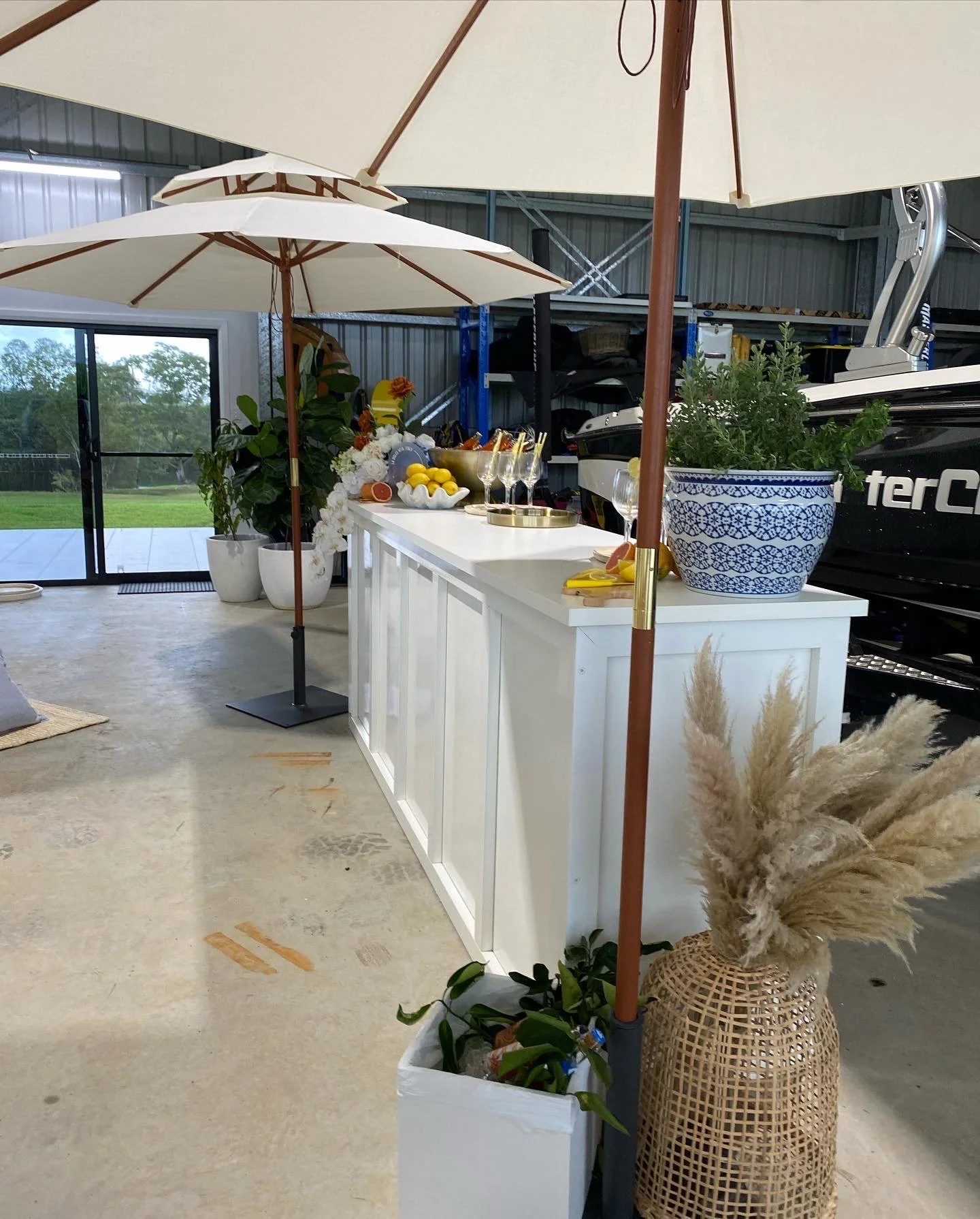 Indoor outdoor party setup with white bar counter, large umbrellas, potted plants, flowers, and a boat in background.